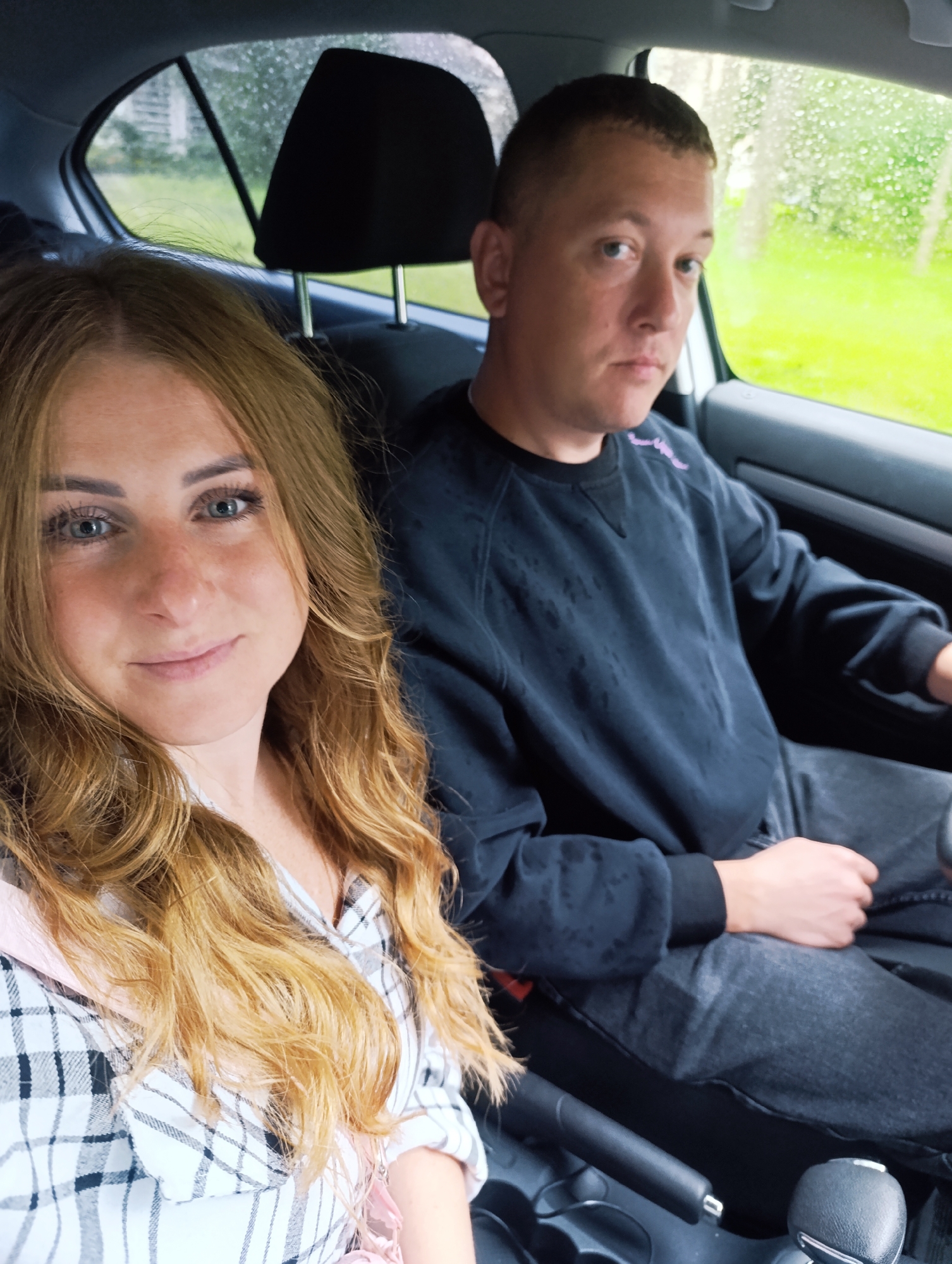 A woman with blonde, wavy hair and blue eyes is in the front passenger seat of a car. She is wearing a white and black plaid shirt. Next to her in the driver's seat is a man with short brown hair and blue eyes. He is wearing a black sweatshirt and dark jeans. The car appears to be parked, and rain is visible on the window. The background shows a green, grassy area and trees. The image was taken in Chernogolovka, Russia.