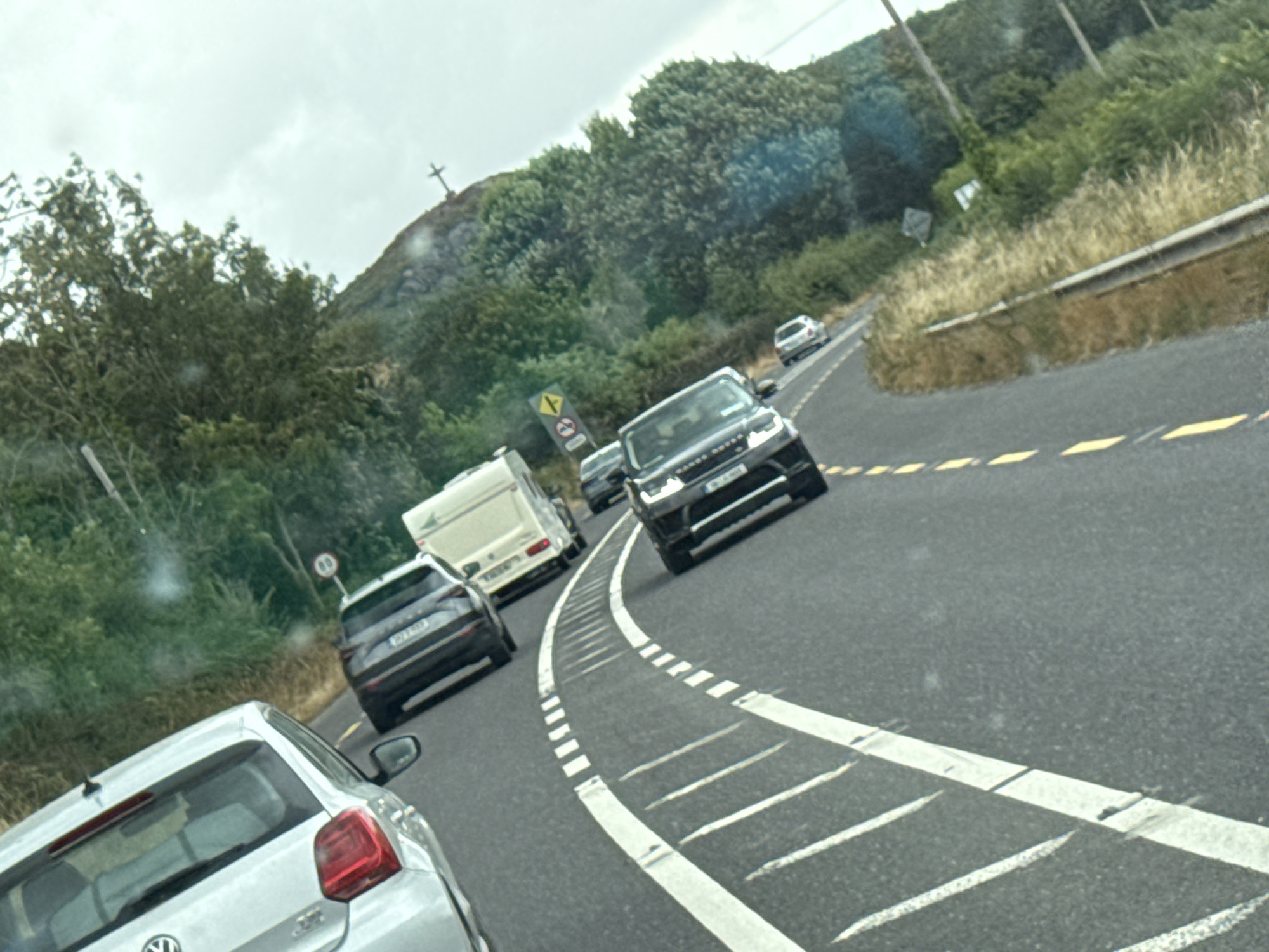 The image depicts a traffic scene on a two-lane road in Ireland, likely on a cloudy day. The road has visible white lane markings, including a curved solid white line and dashed white lines. Several vehicles are present, including a white car on the left, a dark SUV, and a white van. In the distance, another dark SUV and a smaller white car are visible. The road is bordered by a grassy verge on the right, with trees and dense foliage forming a backdrop. Overhead power lines are also visible. The overall impression is one of moderate traffic congestion on a rural or suburban road.