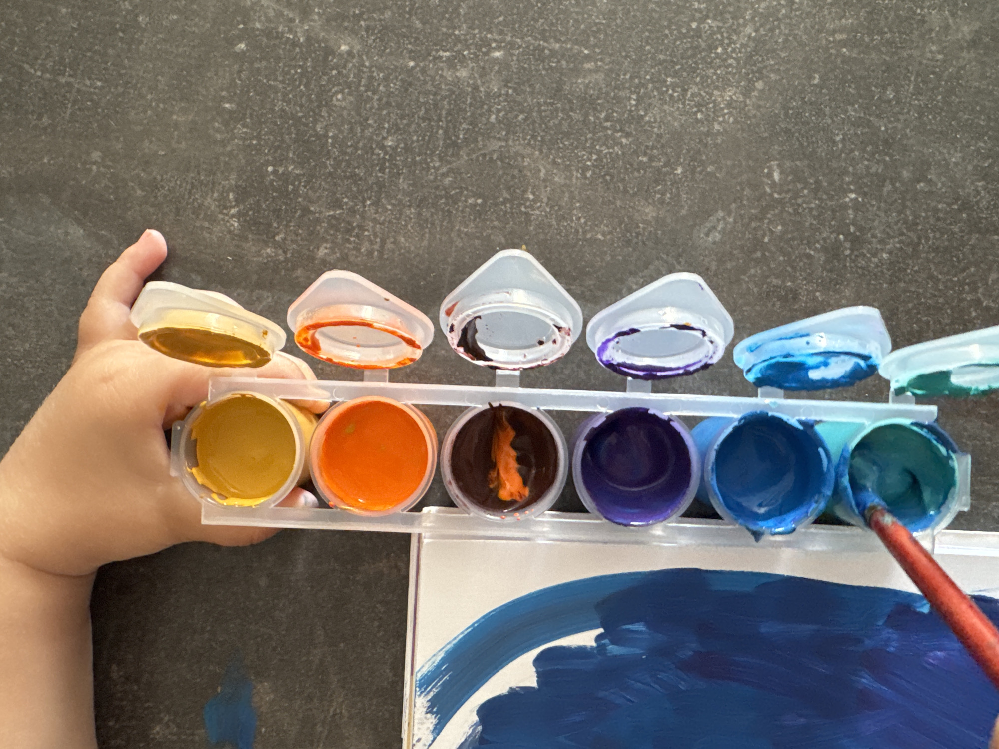 A close-up, overhead shot captures a child's hand reaching for a palette of watercolors, positioned on a dark, speckled surface. The child's skin is lightly tanned, and their fingers are visible as they grasp a small, clear plastic tray containing eight individual wells of paint. The paints are vibrant, with visible shades of yellow, orange, brown, purple, and various blues and greens. A white canvas, partially covered in broad strokes of dark blue paint, is visible to the left of the paint palette. A red-handled paintbrush is dipped into a well of teal-colored paint on the bottom of the palette, suggesting the child is in the process of painting. The background is out of focus, but appears to be a dark countertop. The image does not contain any information that would allow for the identification of its location in Mikhaylovsk, Russia.