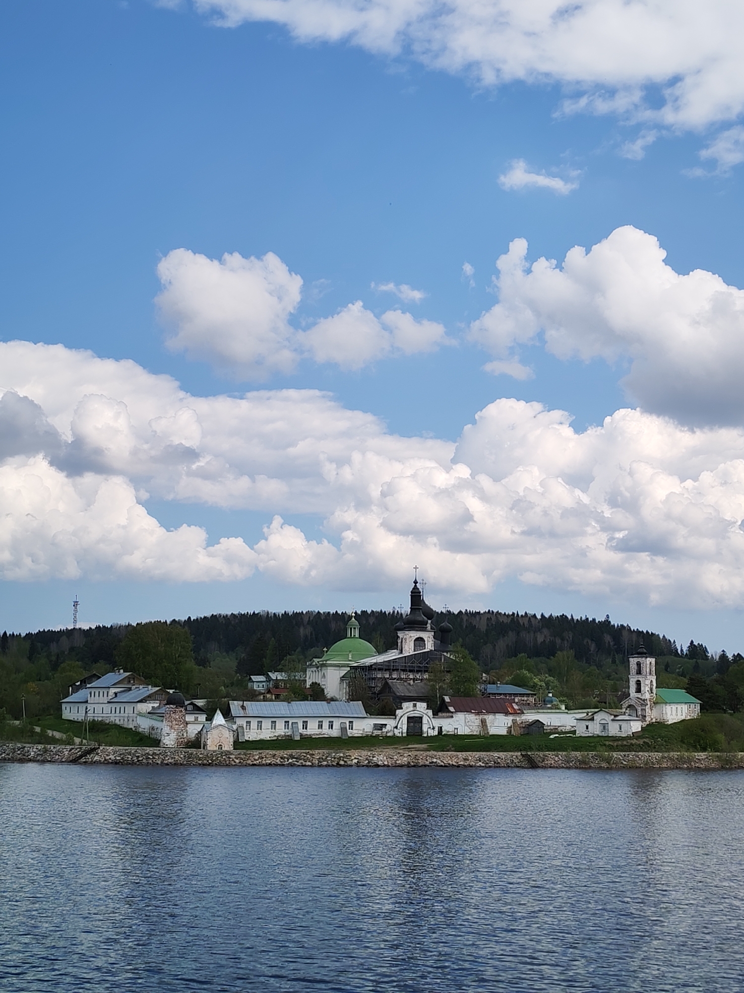 The image shows the Kirillo-Belozersky Monastery in Goritsy, Russia, from across a body of water. The monastery buildings, with white walls and green and black domes, are situated on a low-lying strip of land. Behind the monastery, a dense forest line is visible. The sky is blue with puffy white clouds. The water is a dark blue color.