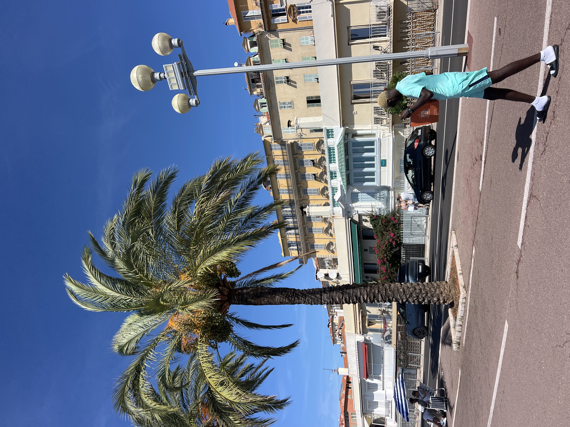 The image depicts a sunny day along the Promenade des Anglais in Nice, France. A prominent palm tree with developing fruit stands in the foreground, casting a shadow on the pedestrian walkway. In the background, a row of multi-story buildings with yellow and cream facades lines the street, featuring balconies and shuttered windows. A black car and a blue SUV are parked on the street, and a lamppost with three spherical lights stands tall. A person with blond hair, wearing a teal t-shirt and shorts, walks across the street carrying an orange bag. Two individuals are seated under a blue and white umbrella on the left side of the image, near the beach. The scene is set against a clear blue sky.