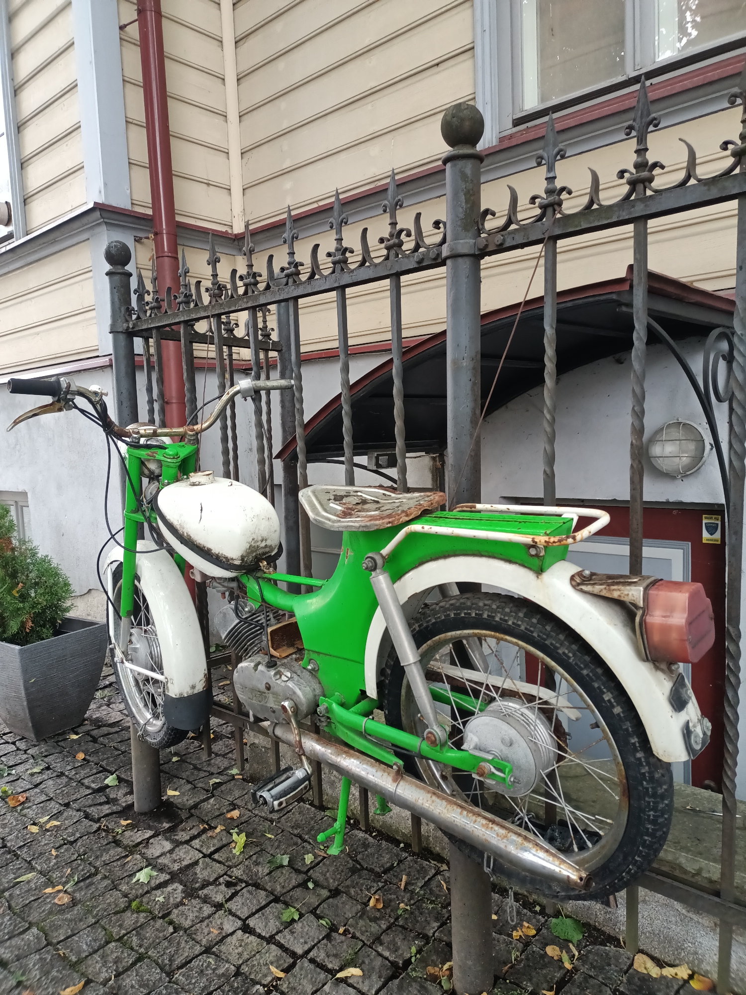 The image shows a vintage green and white moped parked next to a wrought iron fence in Tallinn, Estonia. The moped has a white fuel tank with some visible wear and tear, a green frame and seat, and white fenders. The rear tire is black and has visible tread. The moped is secured to a post by a chain. The background consists of a light-colored building with horizontal siding and a red downspout. The ground is paved with cobblestones and scattered with fallen autumn leaves. A dark gray planter with a small green bush is visible to the left of the moped. A security camera is mounted on the wall behind the fence. The scene suggests an outdoor setting in a residential or historical area of the city.