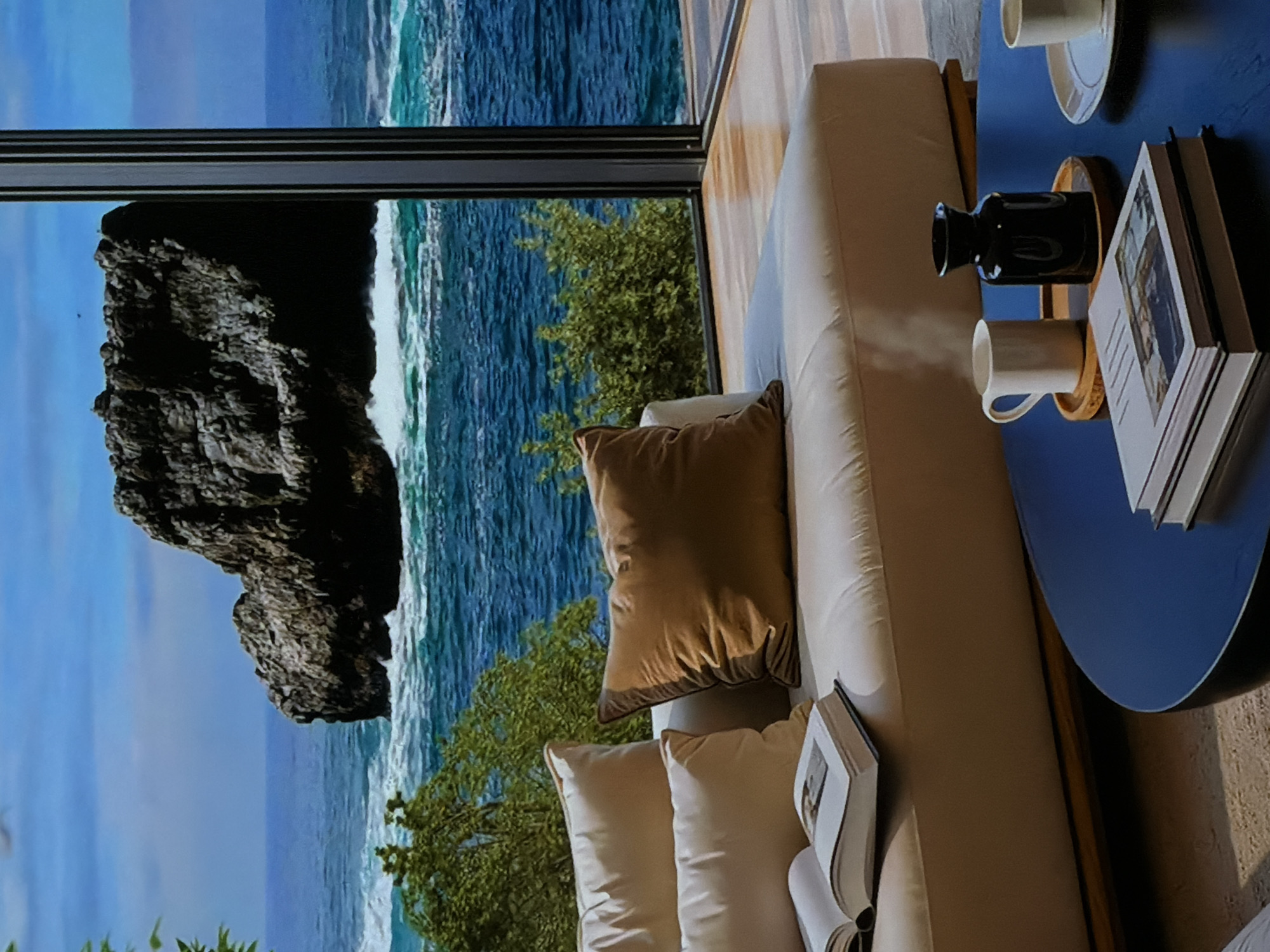 The image presents a serene indoor setting with a panoramic view of a dramatic coastal landscape. On the right, a comfortable, light-colored sofa is furnished with several pillows, including one large brown cushion and three smaller, lighter-toned ones. Adjacent to the sofa, a round, dark blue coffee table holds a black decorative vase, two white mugs (one on a wooden coaster, another on a white saucer), and a stack of books. An open book on top displays an image on its left page, while text on its right page and the spines of other books is illegible. The interior features light-toned wooden elements and a light-colored floor covering.
A large window dominates the left side of the frame, offering an expansive view of a rugged coastline under a clear blue sky. A massive, dark, jagged rock formation or cliff is prominent in the outdoor scene, surrounded by vibrant green trees and bushes. Beyond the land, the vast blue ocean stretches out, with white-capped waves actively crashing.
No people are visible in the image. The scene suggests a peaceful and luxurious retreat designed to maximize the breathtaking sea view. The clear skies and bright illumination indicate it is daytime. The overall impression is one of tranquility and natural beauty.