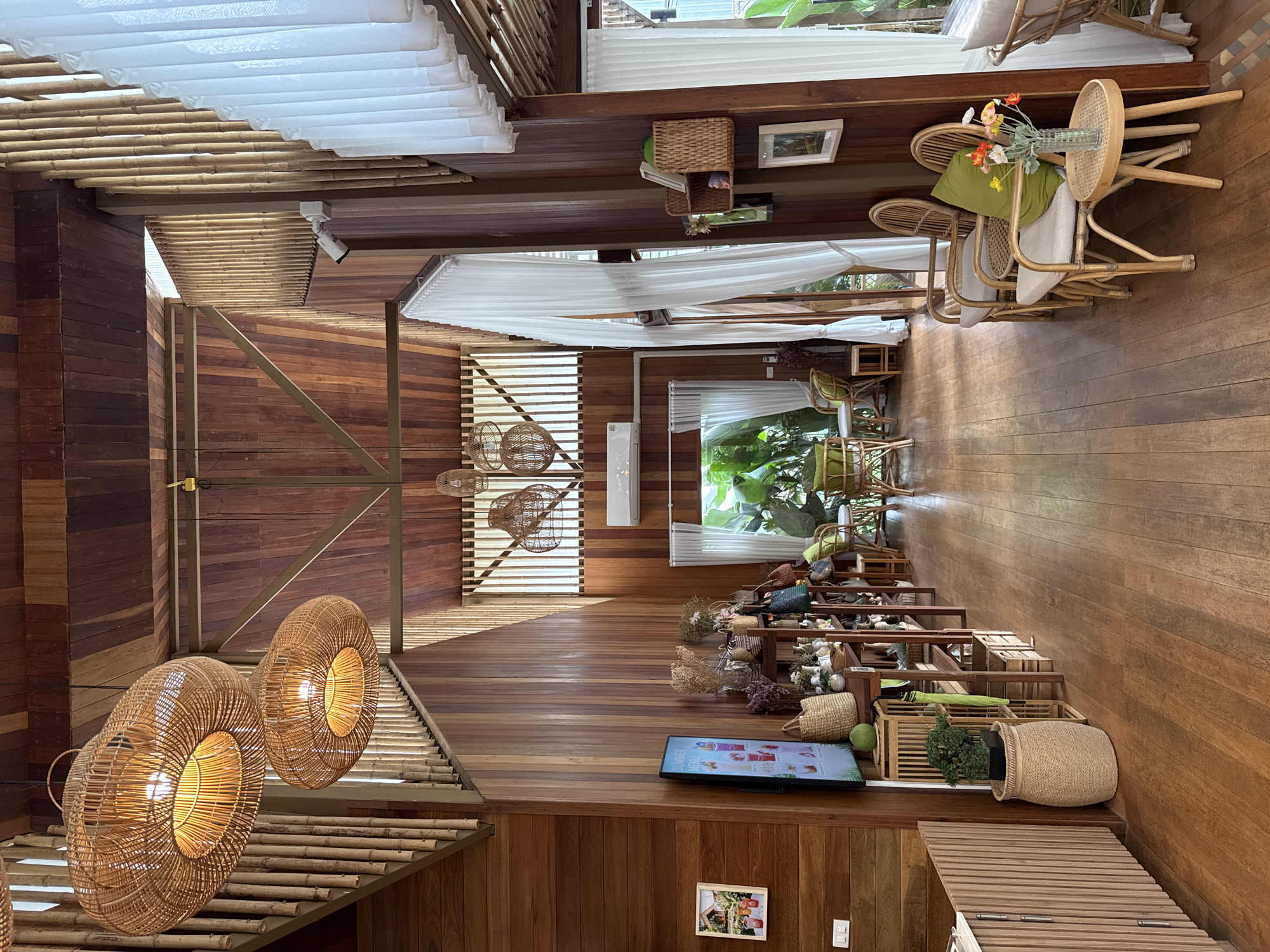 The image depicts the interior of a cafe with a high, vaulted wooden ceiling and walls paneled with light-colored wood. Large, woven rattan pendant lights hang from the ceiling, illuminating the space. A staircase with bamboo railings is visible on the left, leading to an upper level. The floor is made of polished wooden planks.
In the foreground, to the left, is a counter with a fluted wooden front. A flat-screen television mounted on the wall displays a menu with images of drinks. Below the TV, a display shelf holds various items, including woven baskets, plants in pots, and decorative objects. A large, woven basket sits on the floor to the left of the shelf.
The middle ground features a row of woven rattan chairs with light-colored cushions and small round wooden tables. A vase of colorful flowers sits on one of the tables. White sheer curtains hang by large windows, revealing a glimpse of lush greenery outside.
The background shows more of the cafe's seating area and the extensive use of wood and natural materials, creating a warm and inviting atmosphere. The overall aesthetic is rustic and tropical. The image appears to be taken in Sai Ma Subdistrict, Thailand, given the architectural style and surrounding vegetation, typical of cafes in that region. There are no people visible in the scene.