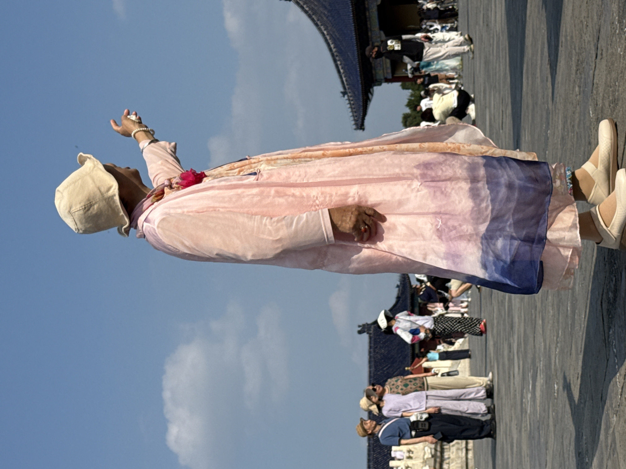 In Dongcheng District, China, a person wearing a wide-brimmed beige hat and a long, flowing pink robe gestures towards the right. The robe has a watercolor-like pattern of blue and purple at the hem and is adorned with a pink floral accessory. The person wears white flat shoes and a pearl bracelet on their left wrist. In the background, several other individuals are visible, some wearing hats and casual attire. To the left, a group of people stands near a white balustrade. To the right, a traditional Chinese building with a dark blue tiled roof is partially visible. The ground is paved with stone, and the sky is clear blue with a few scattered white clouds.