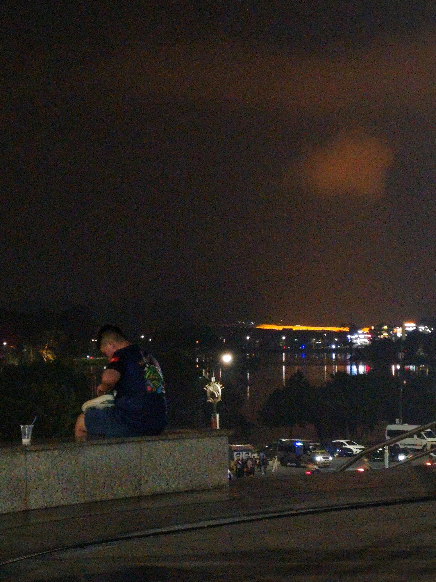 A person is sitting on a stone ledge at night, facing away from the camera. They are wearing a dark blue shirt with a colorful design on the back and blue shorts. A clear plastic cup with a blue straw is on the ledge to the left of the person. In the background, a body of water reflects the lights of a city. Buildings and trees are visible along the water's edge, and a bright orange light emanates from a structure in the distance. Several vehicles, including a white van and a dark SUV, are parked on the right side of the scene. The sky is dark and cloudy, with a subtle orange glow in the upper right corner. This scene appears to be overlooking Xuan Huong Lake in Da Lat, Vietnam, a popular spot for locals and tourists to gather in the evening.
