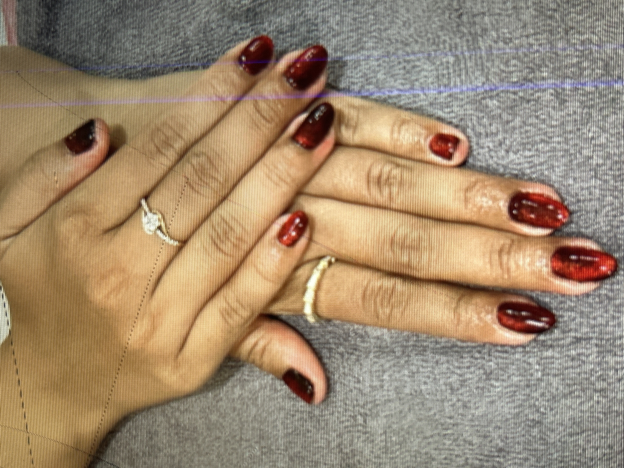 The image shows a close-up view of a pair of hands with dark red, glossy fingernails. The hands are resting on a gray, textured surface. There are two rings visible: a diamond engagement ring on the left hand's ring finger and a delicate, possibly gold or silver, band on the right hand's ring finger. The nails are well-manicured and appear to be artificial, possibly acrylic or gel. The lighting is soft, highlighting the sheen of the nail polish and the details of the rings. There is no indication of the location within Depok, Indonesia, or any specific actions or interactions occurring beyond the display of the hands and jewelry. The overall impression is one of personal grooming and adornment.