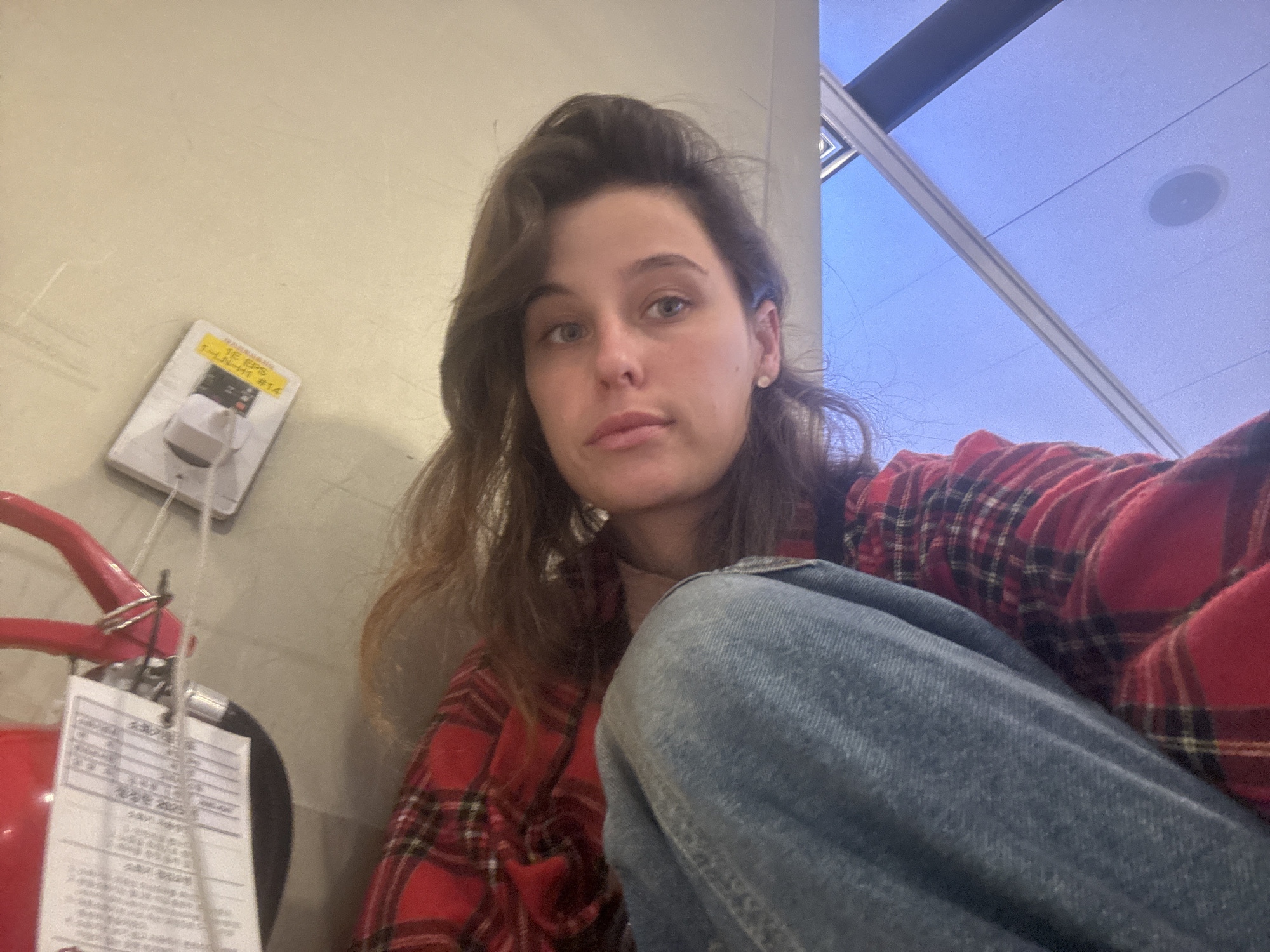 A young woman with brown hair is sitting on the floor in what appears to be an indoor public space. She is wearing a red and black plaid shirt and blue jeans. Her left hand is resting on her right leg, which is bent at the knee. To her left, partially visible, is a red fire extinguisher with a white tag attached. Behind her is a light-colored wall and a large window that looks out onto a bright blue sky. The ceiling is visible through the window, with what appear to be fluorescent lights and a circular speaker. The scene suggests a waiting area or terminal in a building in Seoul, South Korea.