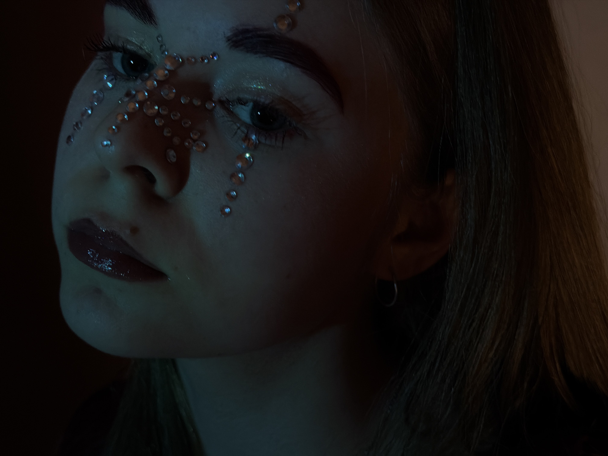 A close-up shot depicts a person's face adorned with numerous small, reflective jewels or beads, arranged in a pattern that extends across their forehead, around their eyes, and down their cheek. The person has dark eyebrows, visible eyelashes, and wears lip gloss. The background is dark, with the right side of the frame showing out-of-focus strands of hair. The lighting is dim and cool-toned, emphasizing the shine of the jewels. The location and city are not discernible from the image.