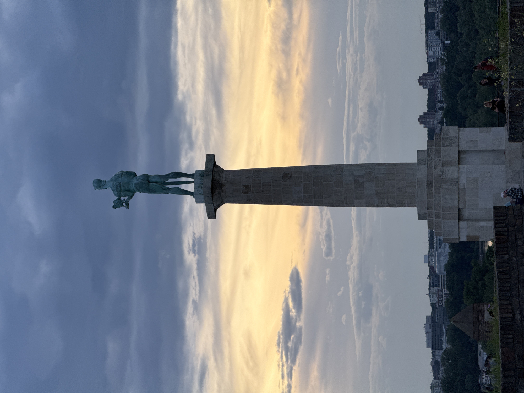 The image shows the Victor Monument in Belgrade, Serbia. The monument consists of a bronze nude male figure holding a falcon, atop a tall stone column. The statue faces left, silhouetted against a dramatic sky with dark blue and golden-hued clouds. Below the column, a stone base structure is visible, with people standing near the edge of the structure. The background reveals urban buildings and green trees, indicating a cityscape. A brick wall is located in the foreground.