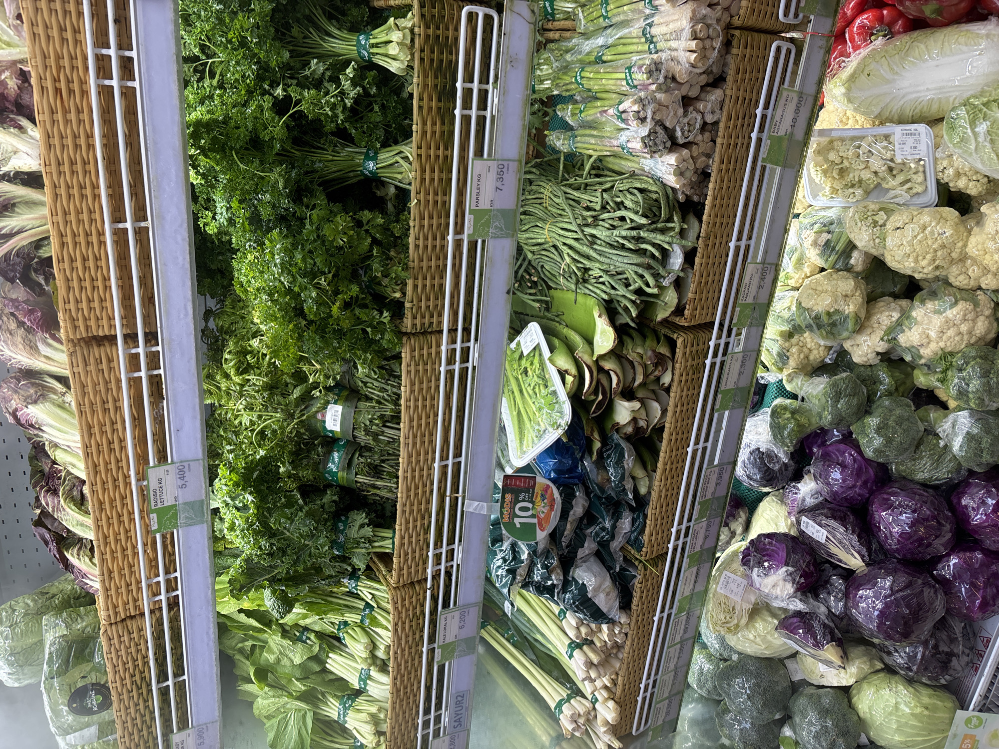 The image shows a vegetable display in a grocery store, potentially located in Kuta Selatan, Indonesia, based on the price tags displaying Indonesian currency (IDR). The shelves are filled with various fresh produce items, including radishes, romaine lettuce, kale, parsley, long beans, baby asparagus, aloe vera, celery, cauliflower, broccoli, and red cabbage. Most items are bundled or wrapped in plastic. Price tags are visible. The vegetables are arranged in wicker baskets.
