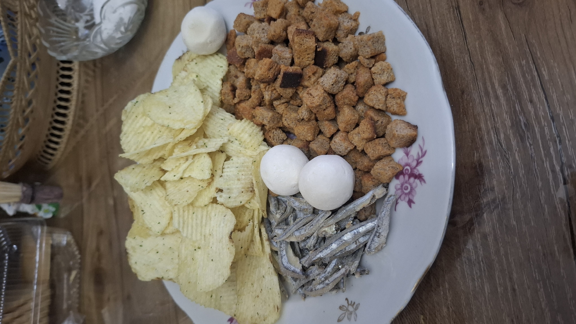 A white ceramic plate with a floral design sits on a wooden table. The plate is filled with a snack assortment, including a pile of ruffled potato chips, a mound of small, square croutons, a portion of dried anchovies, and two white, spherical objects that appear to be small cheese balls or similar. The background shows a blurred environment with wicker baskets and what looks like a plastic container. The lighting suggests an indoor setting. There are no people or animals visible in the image, and no explicit indicators of the location being Shymkent, Kazakhstan.