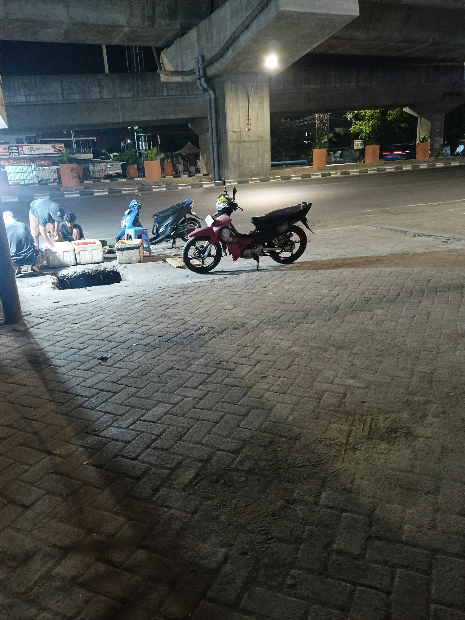 The image depicts a street scene at night under an elevated concrete structure in Makassar, Indonesia. Several motorbikes are parked on a brick-paved sidewalk. In the foreground, three individuals are gathered around what appear to be white styrofoam boxes, with one person appearing to be placing items into a box. To the left, a blue motorbike is parked next to a small blue stool. Further to the right, a red and black motorbike is parked. The elevated structure casts shadows across the scene. A road is visible behind the sidewalk, with some vehicles and streetlights in the distance. Orange planters are placed along the edge of the road. The overall atmosphere is dim, with artificial lighting from streetlights and some illuminated shop fronts in the background.