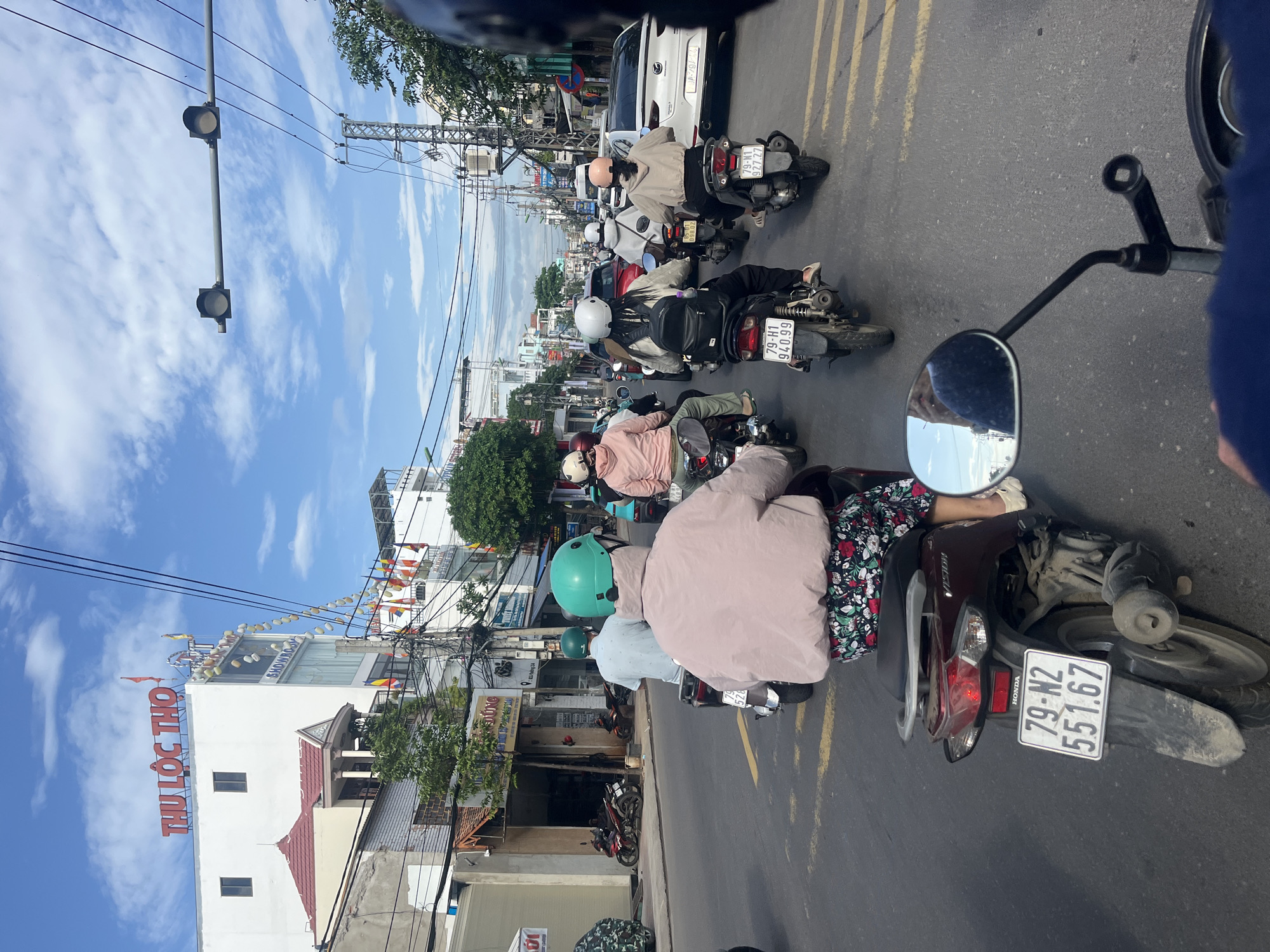 The image captures a vibrant street scene bustling with activity, likely in Bac Nha Trang Ward, Vietnam, as indicated by the location context. The perspective is from a rider's point of view, looking forward into a line of traffic primarily composed of scooters and motorcycles.
Numerous people are visible, mostly wearing helmets and riding scooters. In the immediate foreground, a dark-colored scooter is partially visible, with a person wearing a patterned floral skirt and a light pink jacket. Another person in a light blue helmet and jacket rides ahead. Further down the street, more scooters are lined up, interspersed with a white car in the distance. The street itself has yellow double lines in the center.
The sky is bright blue with scattered white clouds, suggesting a clear day. Buildings line both sides of the street, featuring various architectural styles. Overhead, numerous power lines crisscross the sky, typical of urban areas in the region. Traffic lights are visible suspended from poles against the sky.
Notable details include:
* A "THU LỘC THỌ" sign in red letters on a white building on the left. Below it, a partial "SHOWRO" sign is visible, likely indicating "SHOWROOM."
* Another building has a partially visible "TUNG" sign.
* Several scooter license plates are clear: "79-N2 551.67" (on the foreground scooter, with "HONDA" visible), "79-H1 940.99" (on a black scooter), and "79-N1 927.27" (on a white scooter further down). The number "52" is also partially seen on another building or sign.
* A rearview mirror of the capturing vehicle is visible in the bottom left, reflecting a partial view of the sky and buildings.