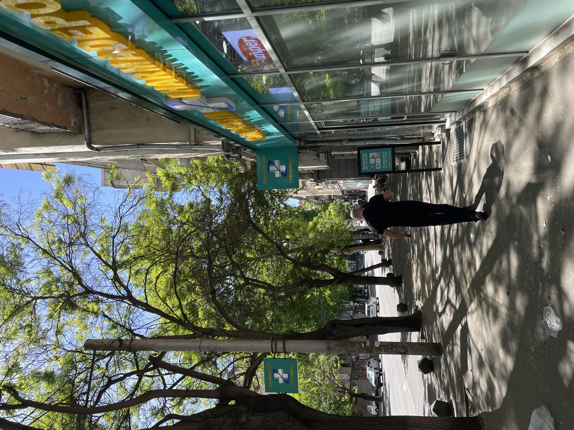 A street scene in Tbilisi, Georgia, shows a man walking along a sidewalk adjacent to a pharmacy. The pharmacy features signage in both Georgian and English, with yellow lettering above. A Pharmadepot sign is also visible. Trees line the street, casting shadows on the sidewalk. Parked cars and buildings can be seen in the background. A streetlamp is also visible.