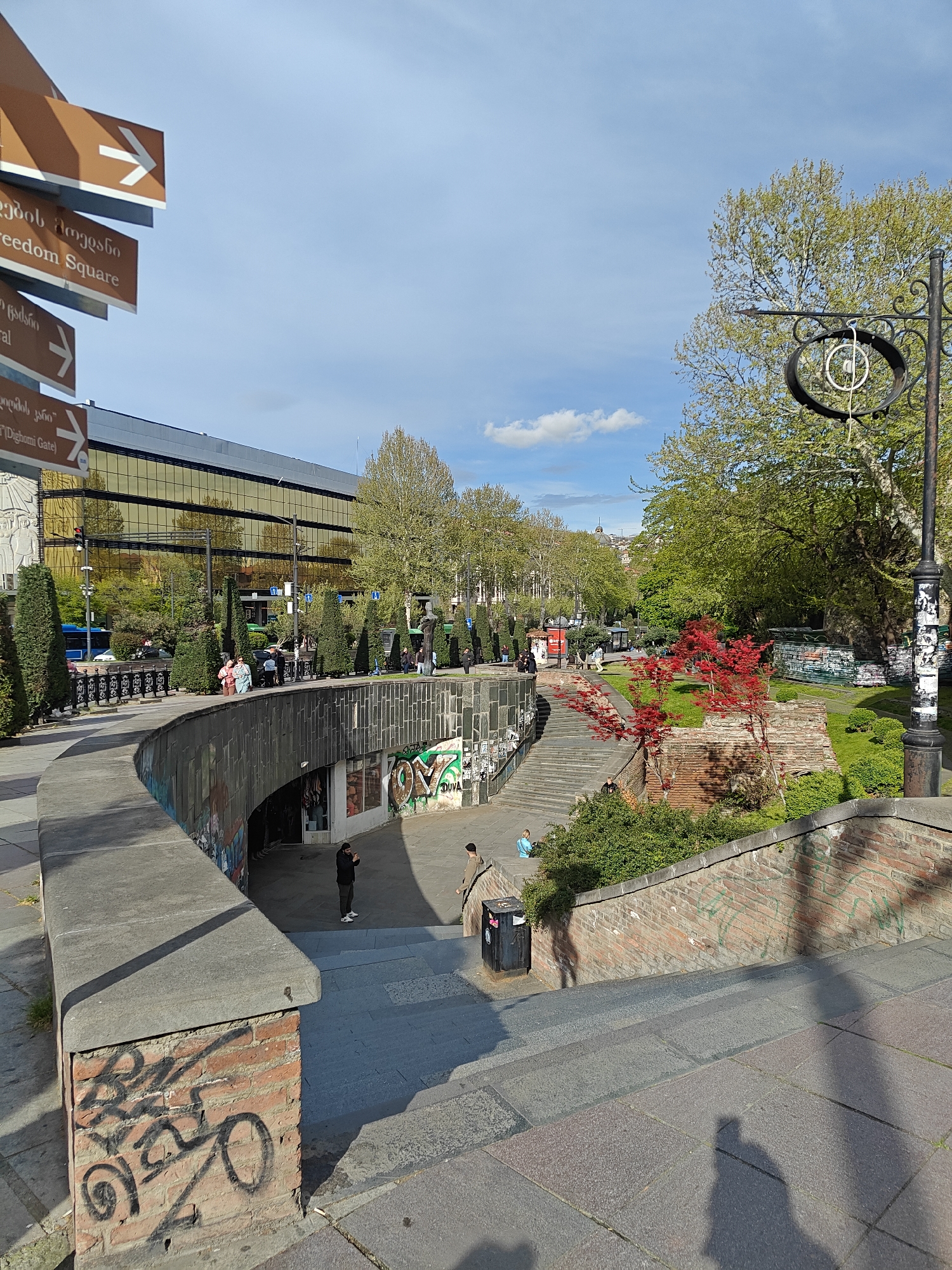 This image captures a vibrant urban scene in Tbilisi, Georgia, on a bright, sunny day, likely mid-morning or late afternoon given the long shadows.
In the foreground on the left, a pedestrian underpass or subway entrance features dark-tiled walls leading down a curved path. The concrete and brick walls surrounding this entrance are covered in various black and green graffiti, including the visible tag "DUVA." A person in dark clothing stands near the underpass entrance.
To the right, a set of stone steps ascends to a landscaped park area. This section is characterized by lush green grass, mature deciduous trees with fresh spring foliage, and trimmed bushes. A small, striking tree with vivid red leaves stands out against the green background. Several individuals are scattered across the steps and the park, some appearing to relax.
A curved pathway runs along the top of the underpass wall, lined with conical evergreen bushes. People are visible strolling along this path. In the background on the left, a large, modern building with reflective golden windows dominates the skyline, mirroring the sky and surrounding environment. A blue bus is partially visible on the street behind the pathway.
Notable objects include a brown directional signpost on the far left, featuring white arrows and text in both Georgian and English. Visible directions include "თავისუფლების მოედანი" (Tavisuplebis Moedani) / "Freedom Square," "ცენტრალი" (Tsentrali) / "Central," and "დიღომის კარიბჭე" (Dighomis Karibche) / "Dighomi Gate." On the far right, an ornate black metal lamp post, also bearing some graffiti at its base, stands tall with a decorative circular element at its top. A black trash can is also visible near the steps.
The sky is clear blue with a few scattered white clouds, contributing to the bright and generally calm atmosphere of the bustling urban environment.