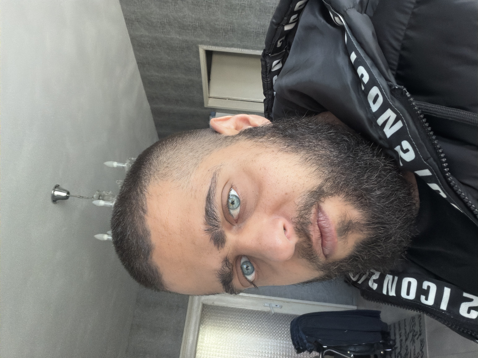 A man with a short haircut and a beard is looking at the camera. He has light blue eyes and fair skin. He is wearing a black jacket with white lettering on the collar. The background is a gray wall with a light fixture and a window or a door. The location is Unknown, Unknown.
