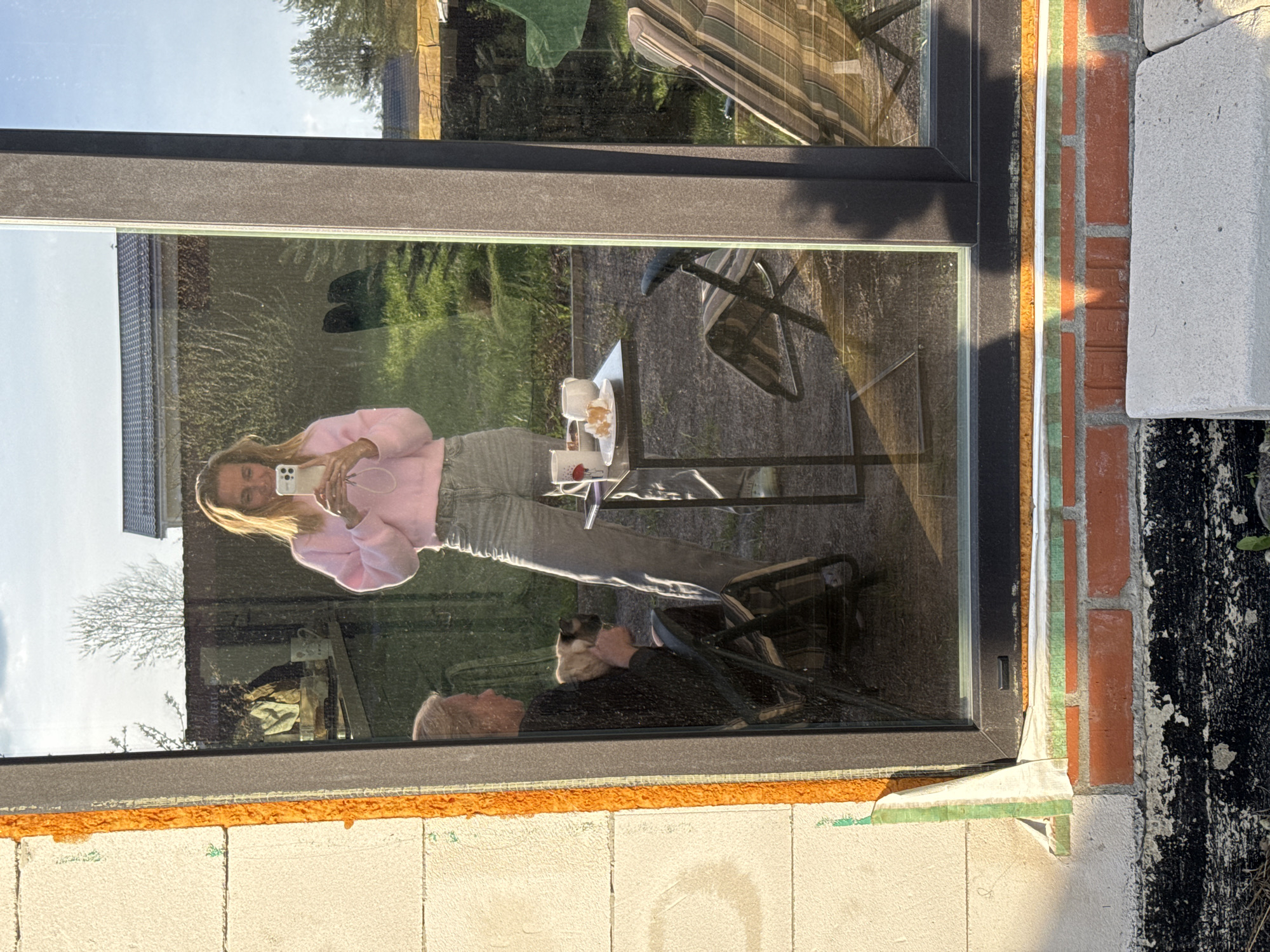 A reflection in a glass window shows a woman with long blonde hair wearing a pink sweater and grey pants holding a smartphone, possibly taking a selfie. Behind her is an outdoor setting with green grass, trees, and a table with cups and a plate. Another person, an older man with white hair, is sitting in a chair, possibly with a dog on his lap. The surrounding area includes construction materials like bricks and white blocks, indicating an ongoing construction or renovation project. The specific location in Steklyannyi, Russia, is not discernible from the image.