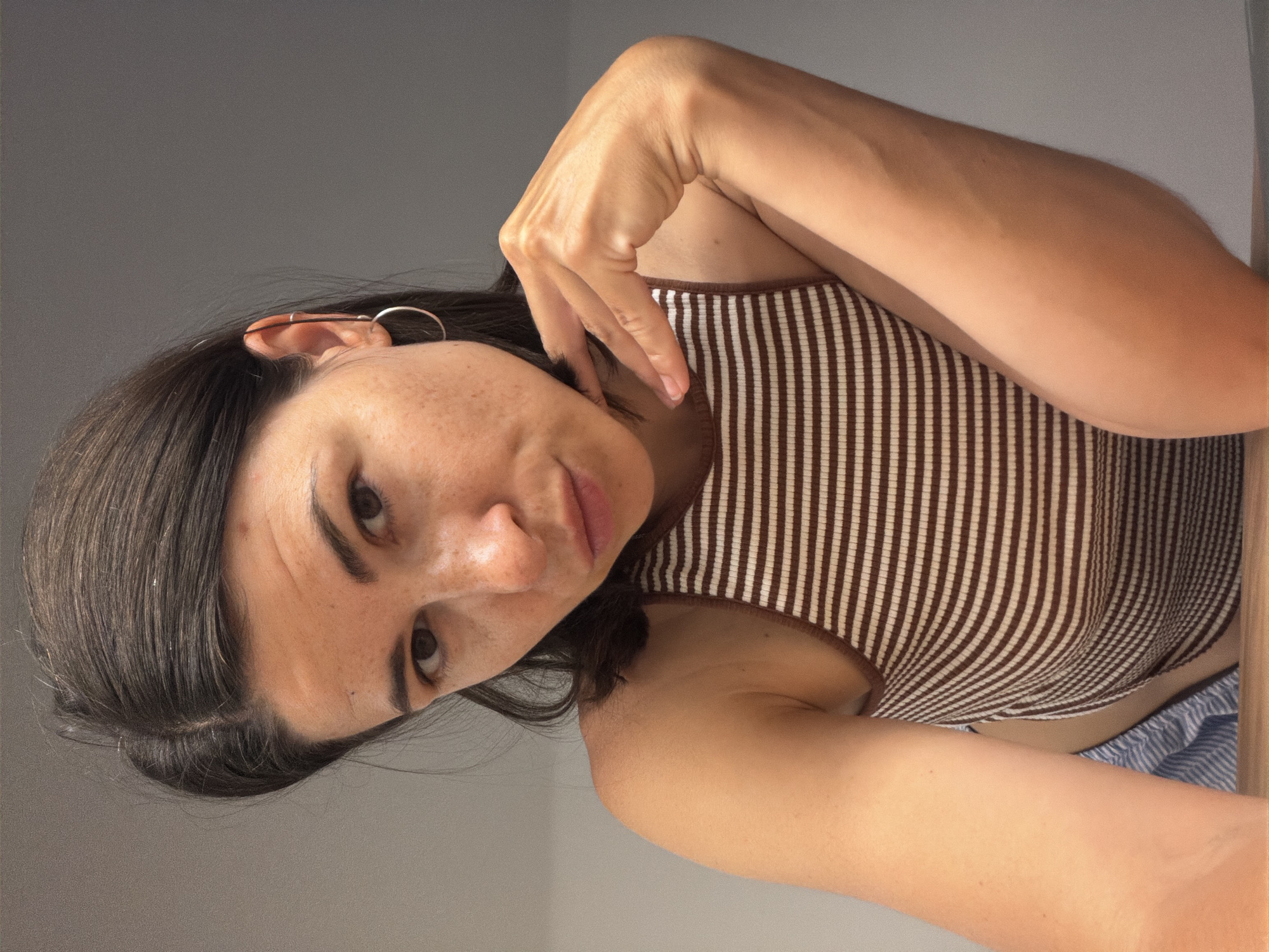 A medium close-up captures a person with fair skin, dark hair, and freckles. She is wearing a brown and white striped tank top and a silver hoop earring in her left ear. Her left hand is raised near her face, with fingers gently touching her cheek. The background is a plain gray wall. No details suggest this image was taken in Budva, Montenegro.