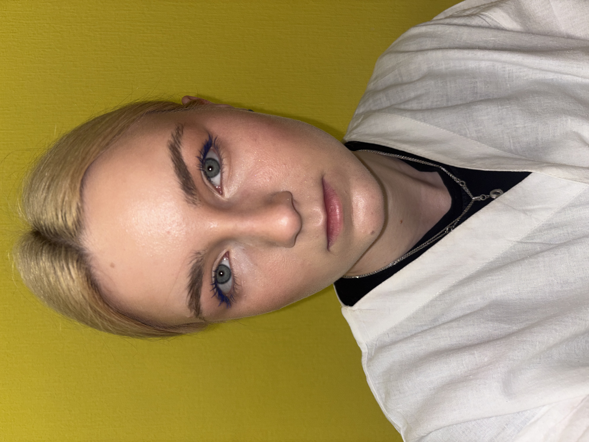 The image displays a close-up, head-and-shoulders portrait of a young Caucasian female with fair skin and blonde hair pulled back. She is wearing a white, open-front garment over a black top, and a silver necklace. Her makeup includes blue eyeliner and blush on her cheeks. The background is a solid, bright yellow wall. There are no discernible elements that would place the location in Sinyavino, Russia.
