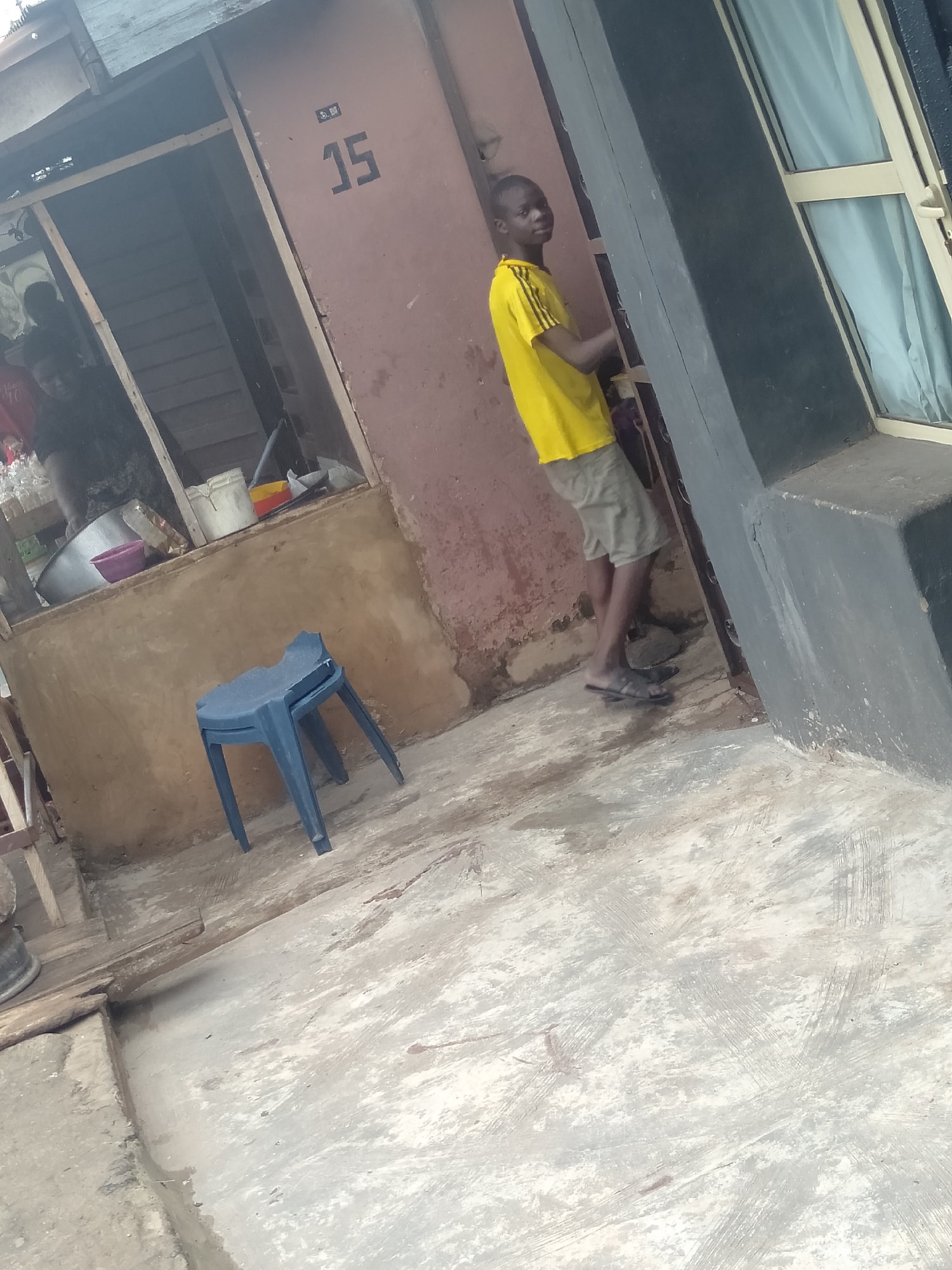 The image depicts a young man standing in a doorway in what appears to be a residential area of Shangisha, Nigeria. The building, marked with the number "15," has a faded exterior and a window with curtains. The man is wearing a yellow t-shirt and khaki shorts. To the left of the doorway, a plastic blue chair sits on a concrete surface. In the background, a person is visible through another doorway or window, near various containers and utensils. The overall impression is a simple, lived-in environment in a developing urban setting.
