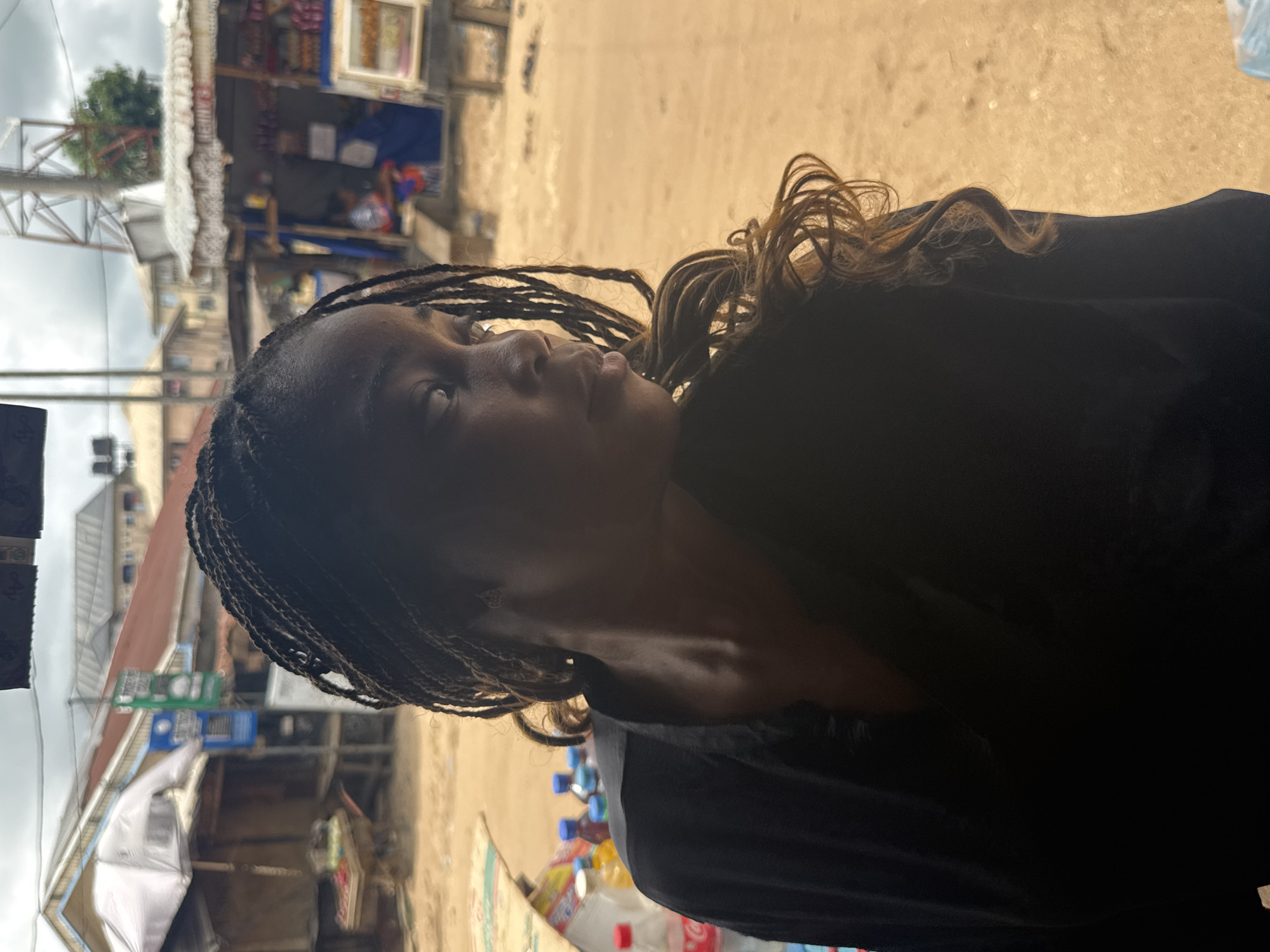 A young woman with braided hair and brown highlights is looking up and to the left. She is wearing a black garment. In the blurred background, there are buildings and structures, some of which are identifiable as small shops or stalls. There are various items for sale visible near the bottom of the frame, including bottled drinks. This scene is most likely occurring in Ihiagwa, Nigeria.