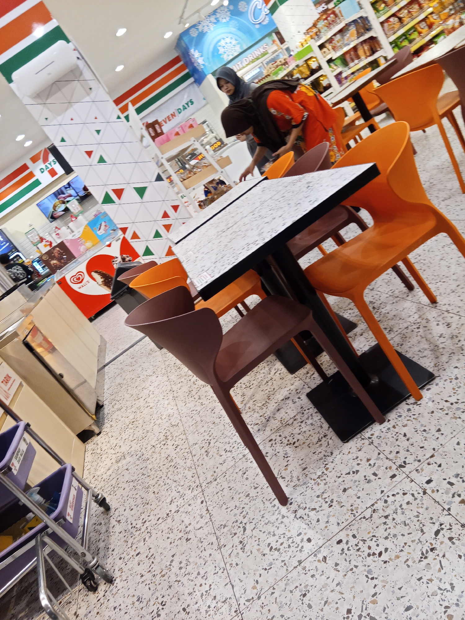 The image depicts the interior of a Seven-Eleven convenience store and cafe area in Lubuklinggau, Indonesia. In the foreground, there are several chairs and a table with a patterned top. Two individuals are visible; one, wearing an orange shirt and headscarf, is bending over the table, possibly interacting with something on it. Another person, also wearing a headscarf, is partially visible to the left. In the background, shelves stocked with various items and refrigeration units are present, consistent with a retail environment. The flooring is a speckled terrazzo tile. The overall lighting suggests an indoor retail space.