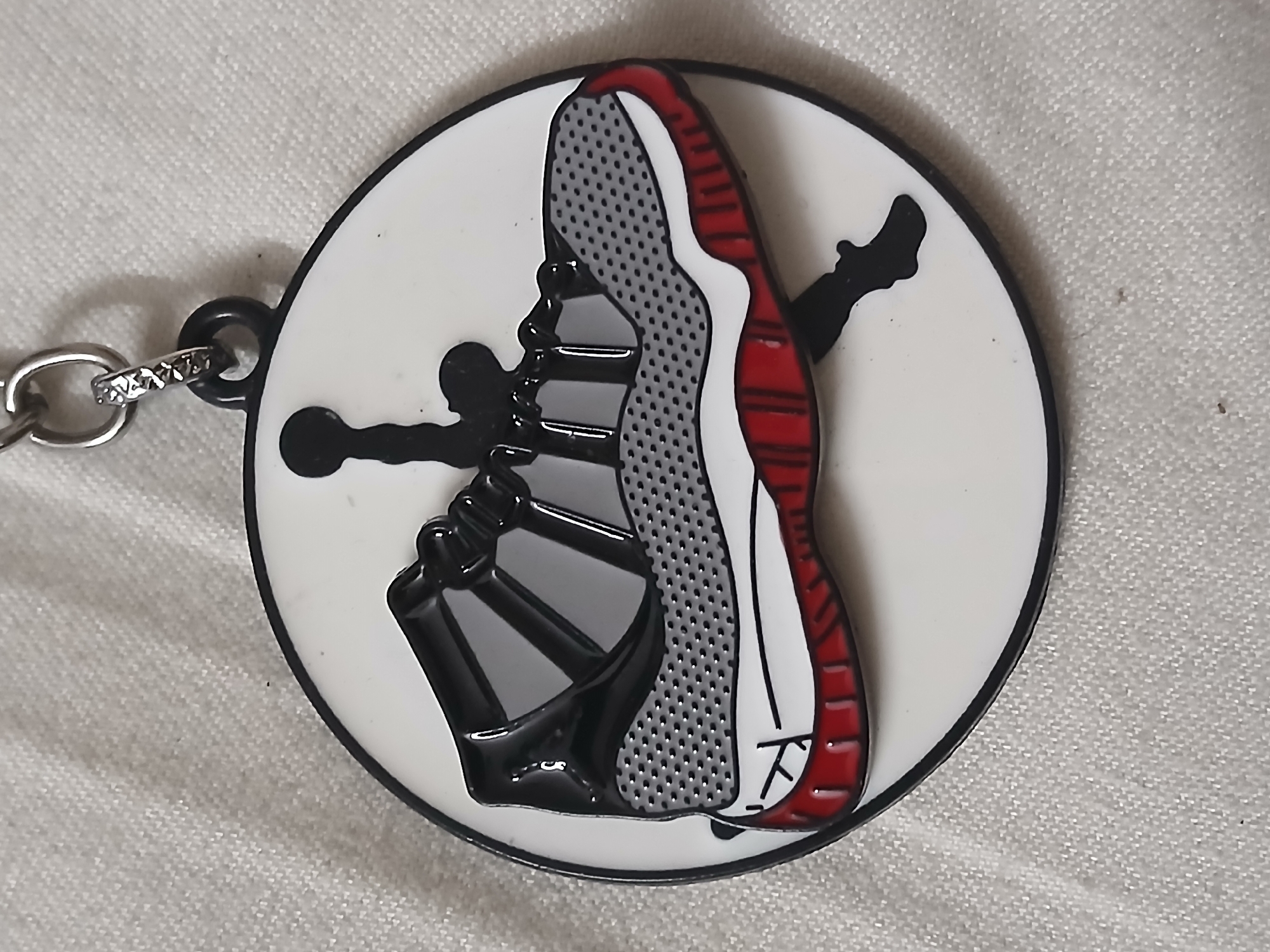 A keychain featuring a partial depiction of an Air Jordan 11 sneaker is displayed on a fabric surface. The keychain is round with a white background and a black border. The sneaker design is in black, grey, red, and white. A black silhouette of the Jumpman logo is also present. The keychain is connected to a silver keyring via a black loop. The fabric surface appears to be light-colored and textured. There is no information about the location or if this photo was taken in Bengaluru, India.