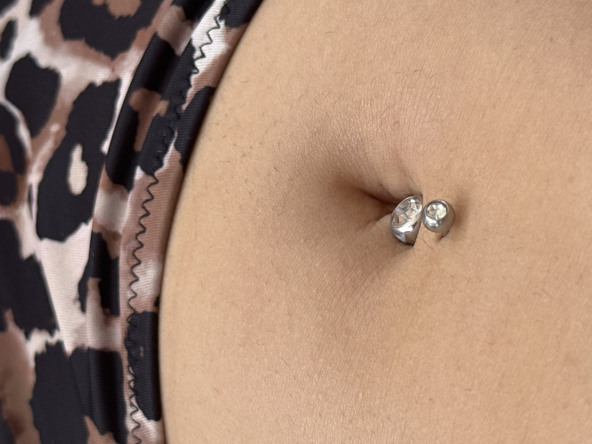 A close-up, low-angle shot focuses on a person's midriff, wearing a leopard-print bikini top. The navel, pierced with a silver belly button ring featuring two clear gemstones, is centrally located. The skin is lightly tanned with visible pores and a few fine hairs. The background is out of focus, showing the patterned fabric of the bikini. The image does not contain information about the city of Limenaria, Greece.