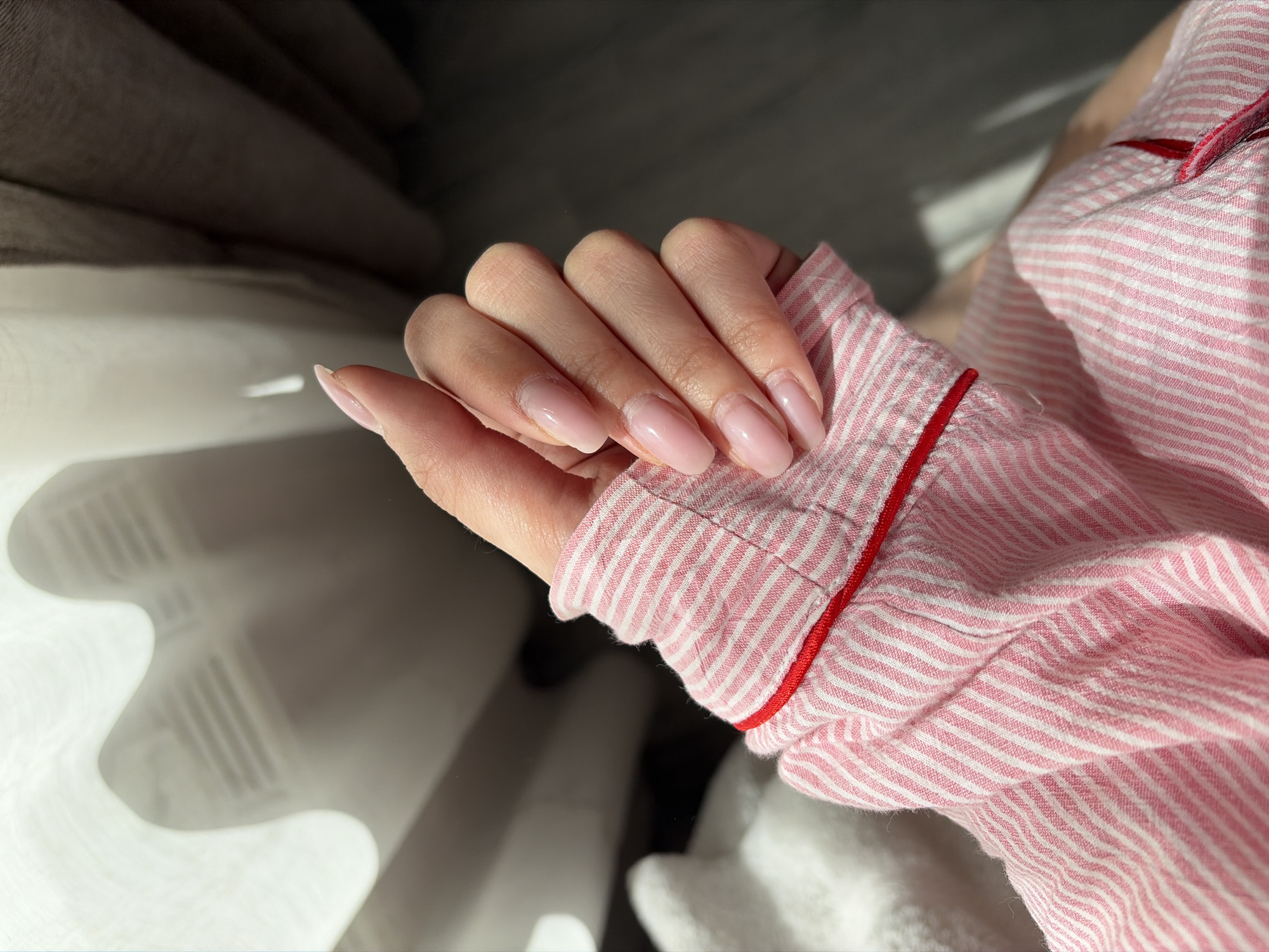 A person's hand rests on a pink and white striped sleeve with red trim. The fingernails are long and painted a light pink color. The background includes what appears to be bedding and a wall with light and shadow. No information indicates that this image was taken in Izhevsk, Russia.