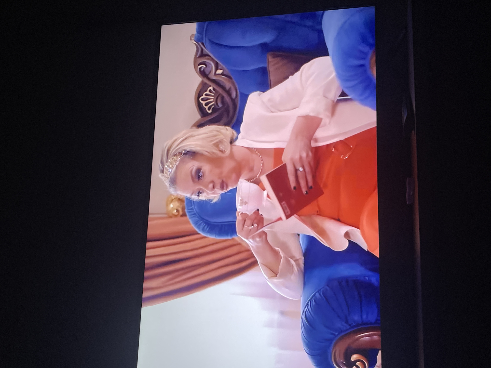 A woman with blonde hair, wearing a white blazer over an orange dress and a sparkly headband, is sitting in a large blue armchair and holding a teacup in her left hand and a red book in her right hand. She is looking to her left. The armchair has carved wooden accents on the arms and back. Behind her, there are draped curtains in a peachy-orange color. The scene appears to be indoors, possibly in a living room or a hotel lobby. There is no identifiable information within the image to indicate that this scene is located in Tallinn, Estonia.