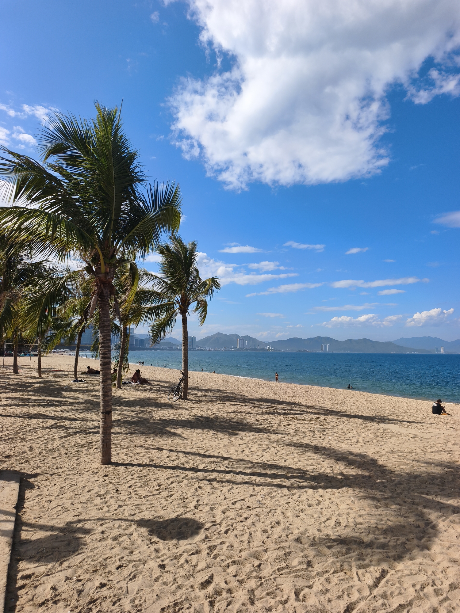 The image depicts a sandy beach in Nha Trang, Vietnam, under a bright blue sky with scattered white clouds. Palm trees are prominently featured in the foreground, casting long shadows across the sand. In the middle ground, several people are relaxing on the beach, some lying down and others sitting. A bicycle is parked near one of the palm trees. The turquoise ocean stretches to the horizon, where mountains and city buildings are visible in the distance. The scene conveys a sense of a leisurely day at the beach.