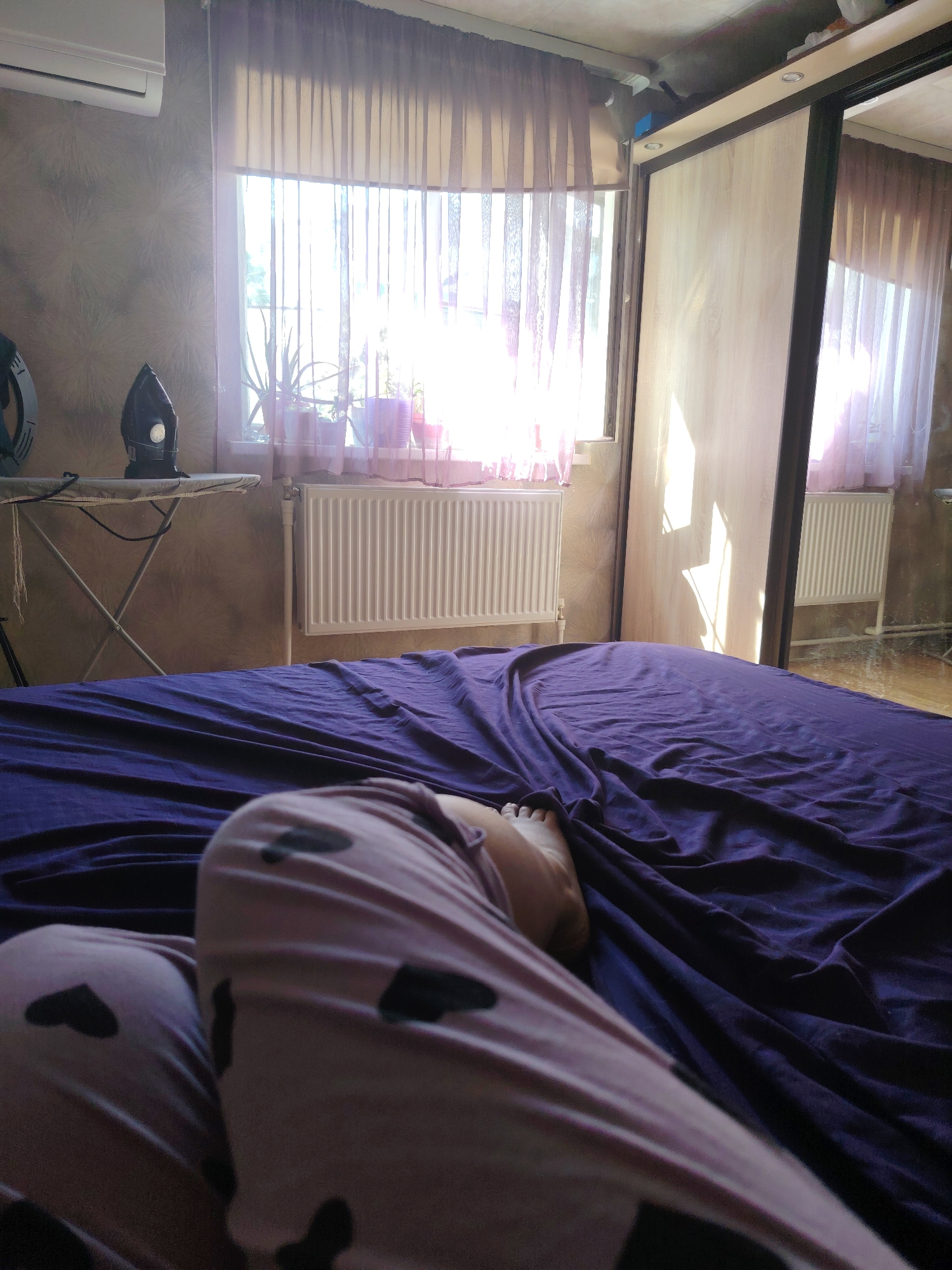 The image shows a bedroom interior with a person lying on a bed. The bed is covered with a dark purple sheet. The person is wearing pink pajamas with black heart patterns. Their left leg is visible, with a foot resting on the sheet. In the background, there is a window with sheer curtains, sunlight streaming through, and plants on the windowsill. An ironing board with an iron is placed to the left of the window. A white radiator is positioned below the windowsill. To the right of the window, a large wardrobe with mirrored doors is visible. The overall lighting suggests it is daytime, with sunlight creating strong highlights and shadows. The room appears to be in a residential setting, possibly an apartment in Rostov-on-Don, Russia, given the presence of a radiator and wardrobe typical of the region.