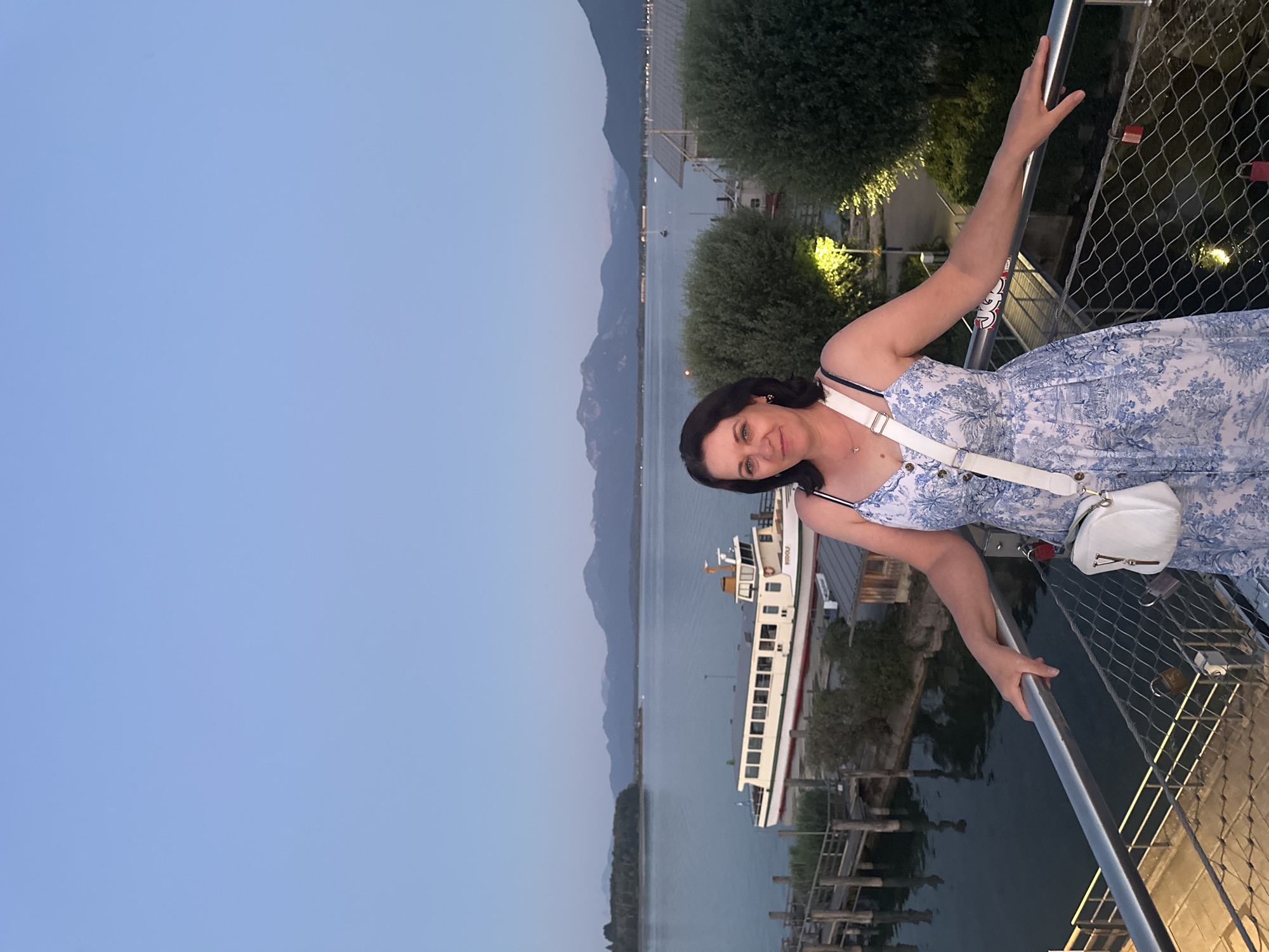 A dark-haired woman in a blue and white patterned dress and a white crossbody bag stands on a metal walkway, leaning on the railing. The walkway overlooks a body of water, where a large white boat with green trim is docked. In the background, a line of trees separates the water from mountains under a twilight sky. The scene is set in Prien am Chiemsee, Germany, possibly at the harbor.