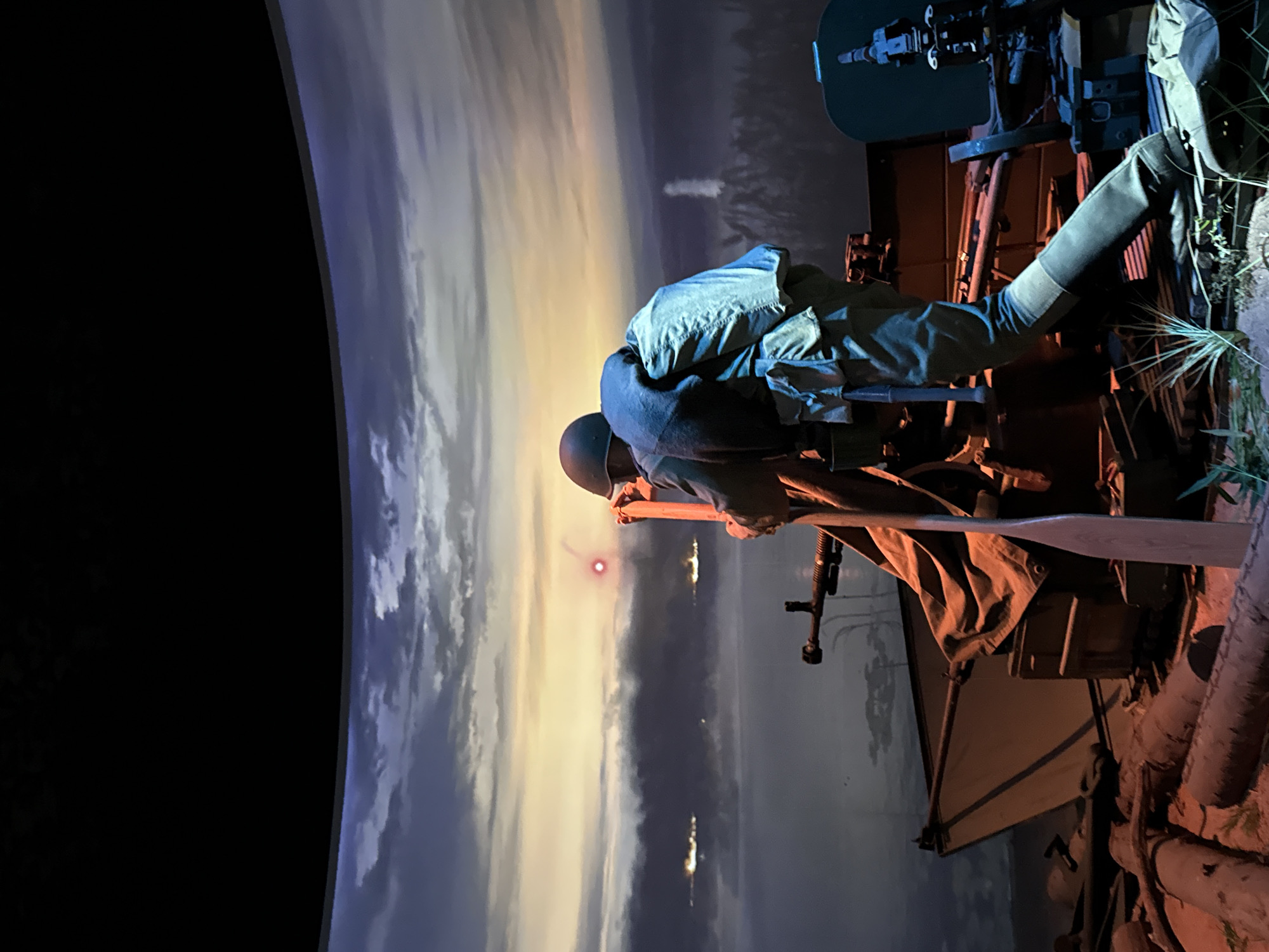 This is a medium shot of a diorama, likely a museum exhibit, depicting a military scene at dusk or dawn. In the foreground, a mannequin dressed in military attire, including a helmet, uniform, and backpack, is positioned behind a machine gun. The mannequin is hunched over, appearing to aim the weapon. The machine gun is mounted on a wooden structure and partially covered by a canvas tarp. In the background, a panoramic painted backdrop simulates a body of water with distant lights and a sky transitioning from dark to a warm, hazy light, possibly representing the sun. The overall scene evokes a wartime setting, possibly related to coastal defense or naval warfare. The scene is likely located in the city of Zelenaya Roshcha, Russia, given its specific location within the city.