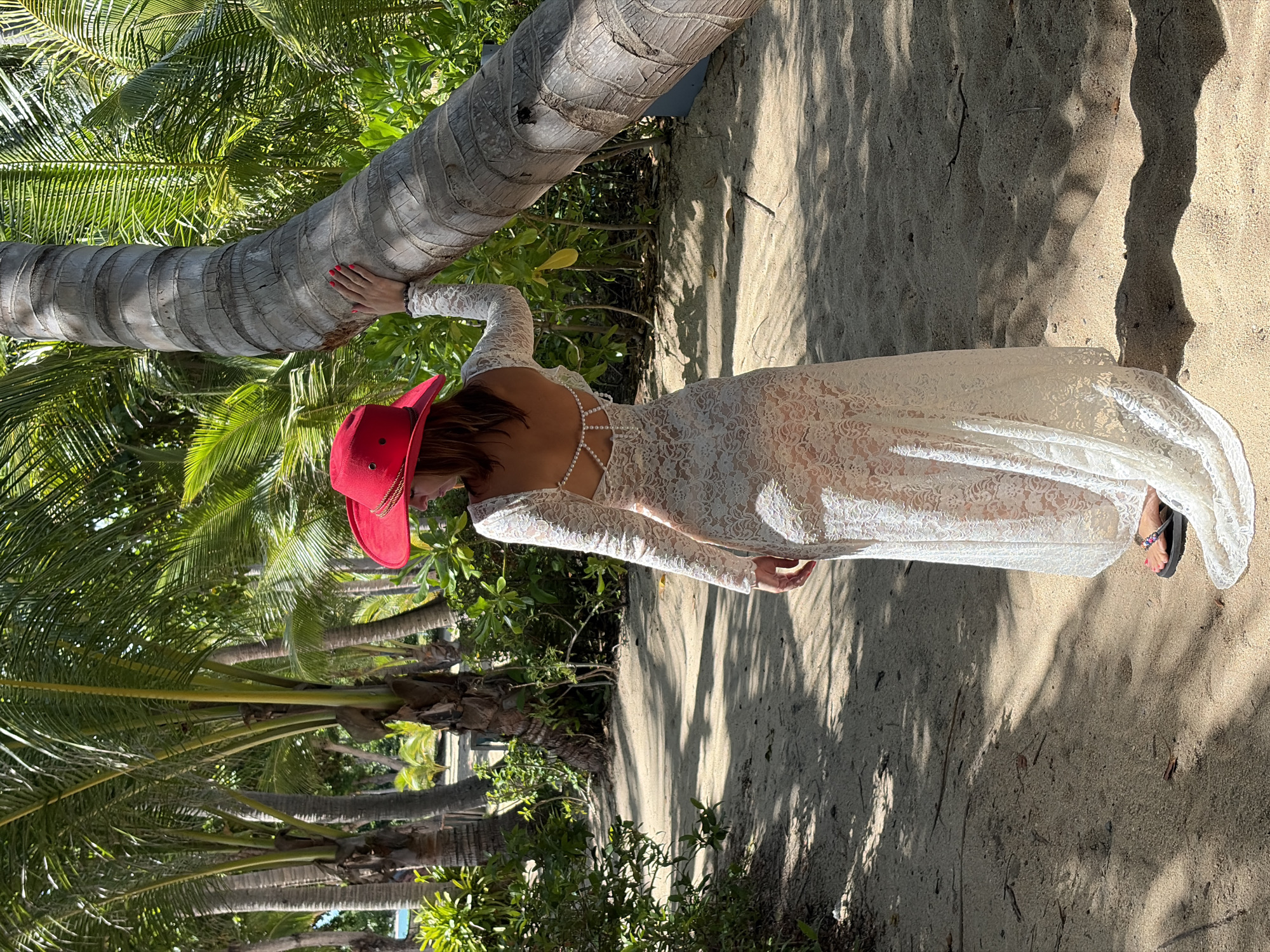 A woman wearing a white lace dress and a red hat stands on a sandy path under palm trees. She is leaning against a large palm tree, with her right hand resting on the trunk. The dress has long sleeves and an open back adorned with pearls. The path is lined with lush green vegetation and the ground is covered in sand. The lighting suggests it is daytime, with dappled sunlight filtering through the palm fronds. The location appears to be a tropical beach or park area.