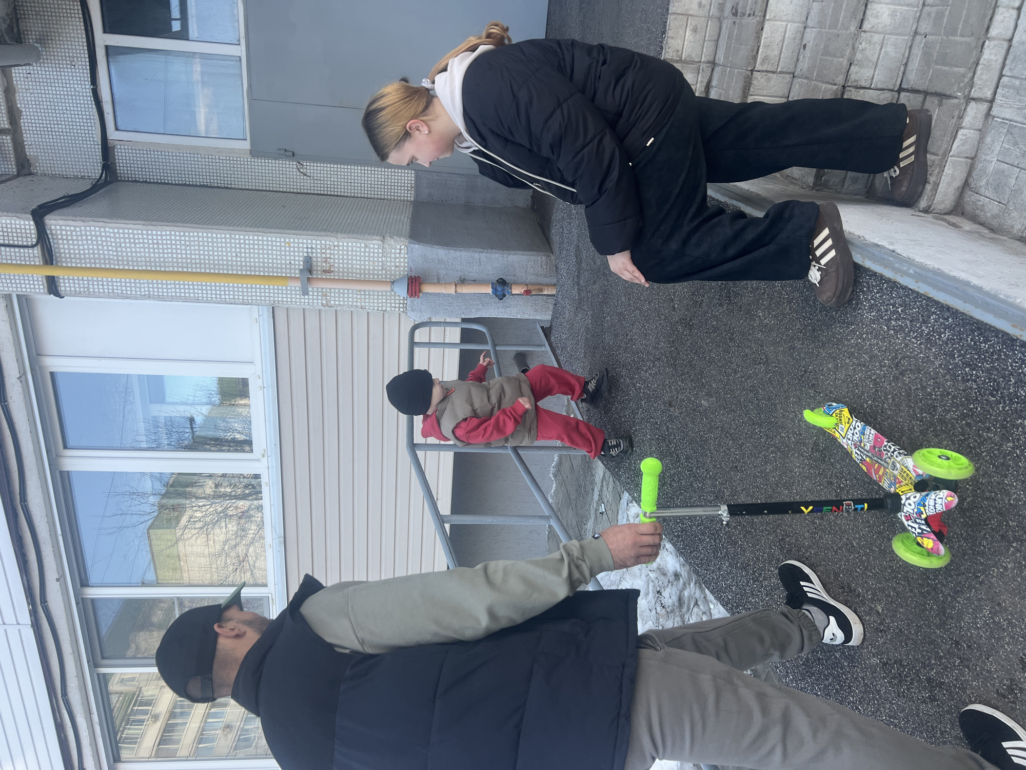 Outside in Chaikovsky, Russia, a woman in a black puffy jacket and brown Adidas sneakers leans forward looking at a young boy in a black beanie, red pants, and brown vest who stands on a set of silver stairs. Beside the stairs is a yellow pipe. A man in a black baseball cap, black vest, light green long sleeve shirt, and gray pants is partially visible. He holds a green scooter. Near him is a scooter with lime green wheels and multicolored decals. A building with two windows is visible in the background. There is snow on the ground. The ground is gray and appears to be concrete.