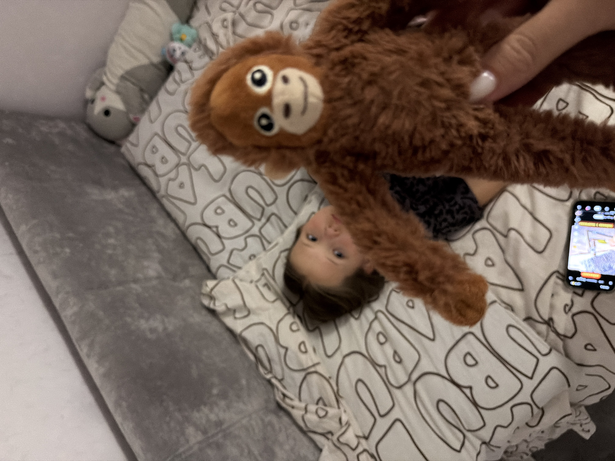 A child lies on a bed in Chaikovsky, Russia. The bed has a gray velvet headboard and a white duvet cover with black stylized letters. A brown stuffed monkey is held above the child. A smartphone with its screen visible sits on the bed. There is a stuffed gray toy animal near the headboard. A hand with white nail polish is holding the monkey.