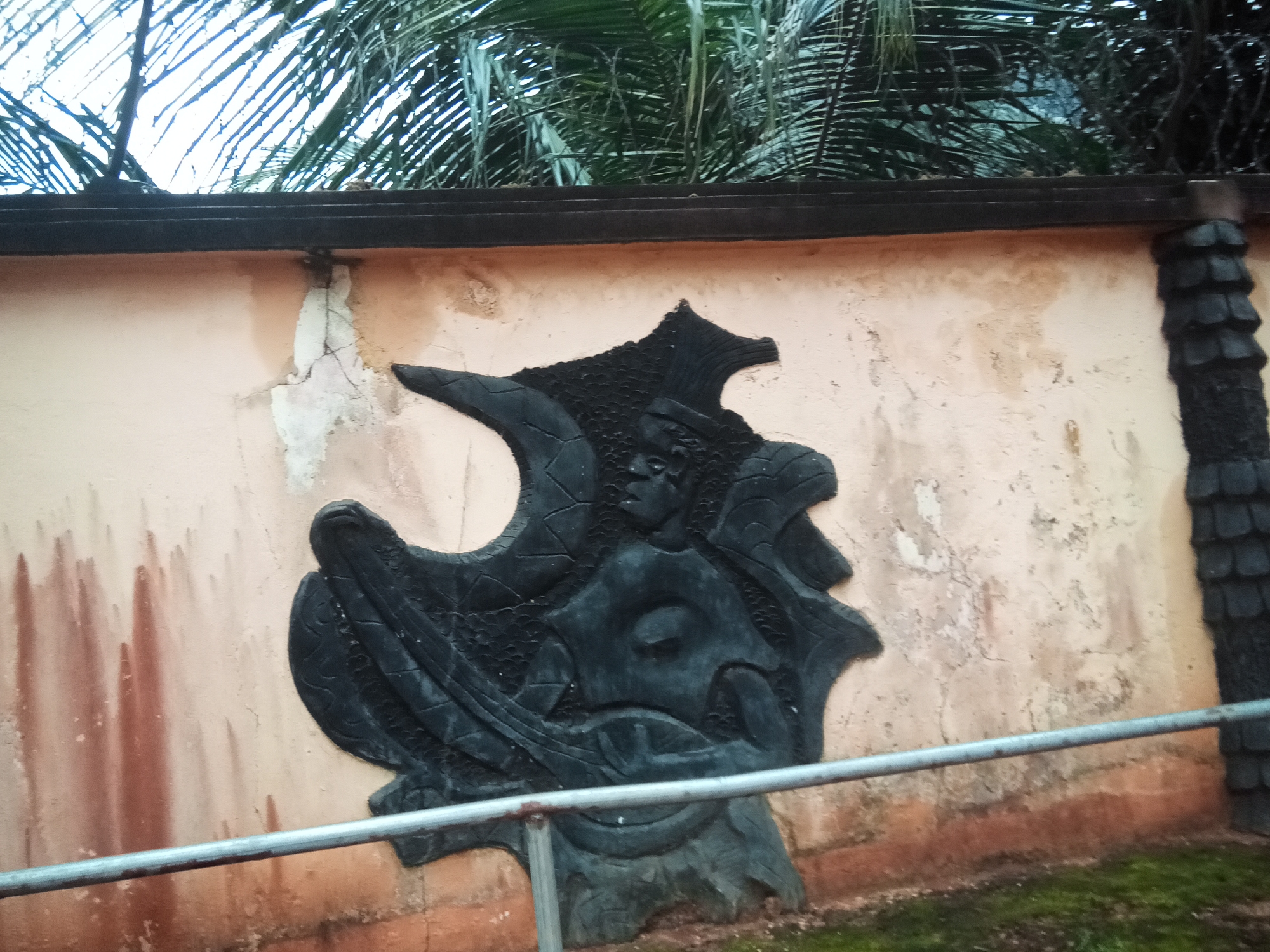 The image shows a close-up view of a textured, dark, abstract sculpture mounted on a light-colored, weathered concrete wall. The sculpture appears to be a stylized depiction of a creature or face with sharp edges and intricate patterns. The wall exhibits signs of age and damage, with cracks and discolored streaks running down its surface. Above the wall, the fronds of palm trees and a dark, barbed wire fence are visible against a bright sky. In the foreground, a metal railing runs horizontally across the frame, with patches of grass and dirt visible below. The scene is outdoors and appears to be in a tropical or subtropical environment. The location is identified as Oko Town, Nigeria.