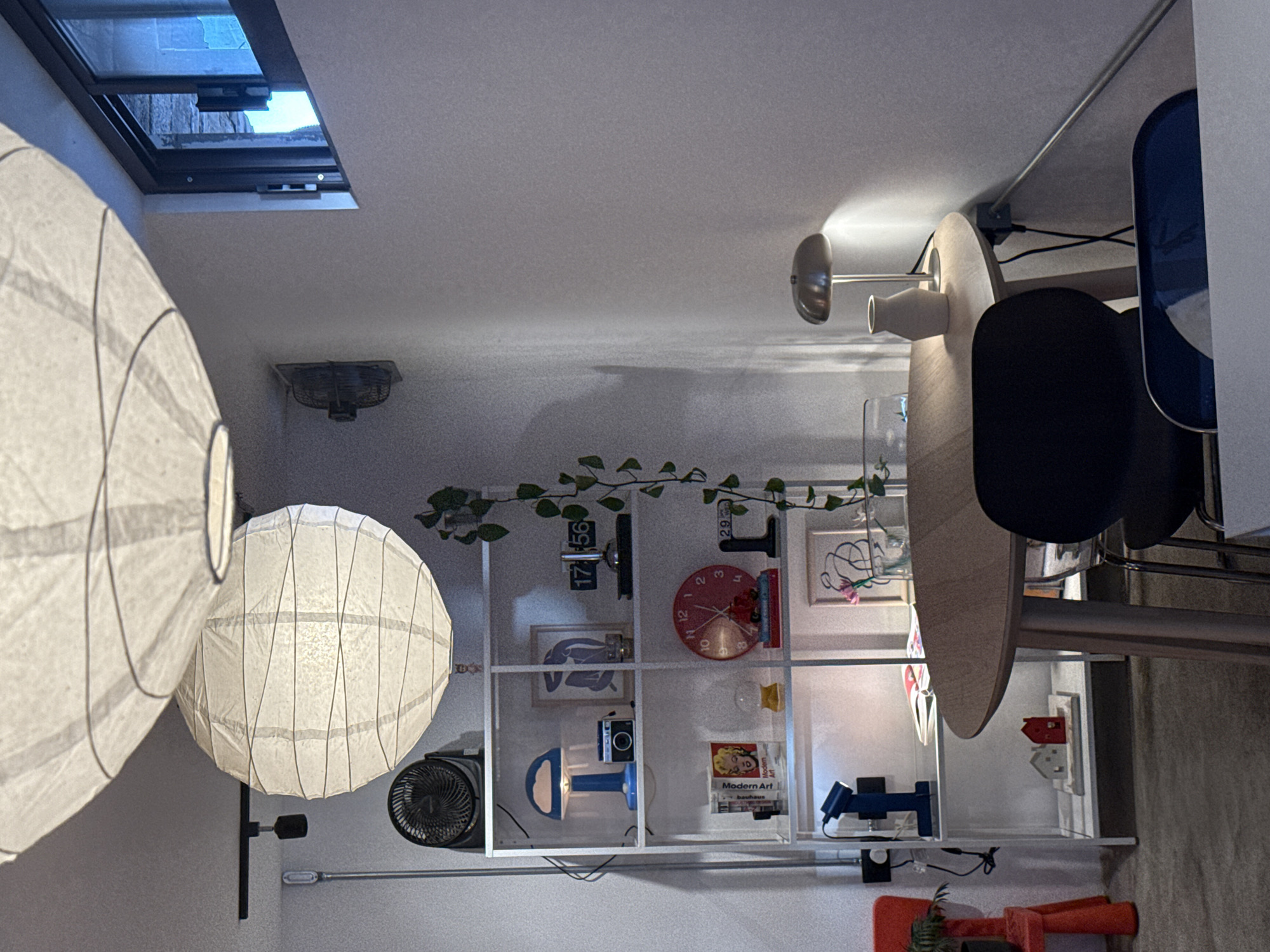 This indoor shot shows a room interior, likely in a house or apartment. The objects in the foreground consist of two white, spherical paper lanterns that hang from the ceiling or wall. A skylight is visible near the top left corner of the frame, letting in natural light.
To the right, there is a round wooden table with a modern lamp attached to it. A chair with a blue back and a black seat is situated next to the table.
In the background, there is a white shelving unit that displays various decorative items, including a red wall clock, a digital clock, a miniature camera, framed artwork, and a variety of small objects. A small desk fan is also on one of the shelves. A few artificial green vines are draped across the top of the shelving unit.
The exact location in the city of San Andrés Cholula, Mexico, cannot be determined from the information in this image.
