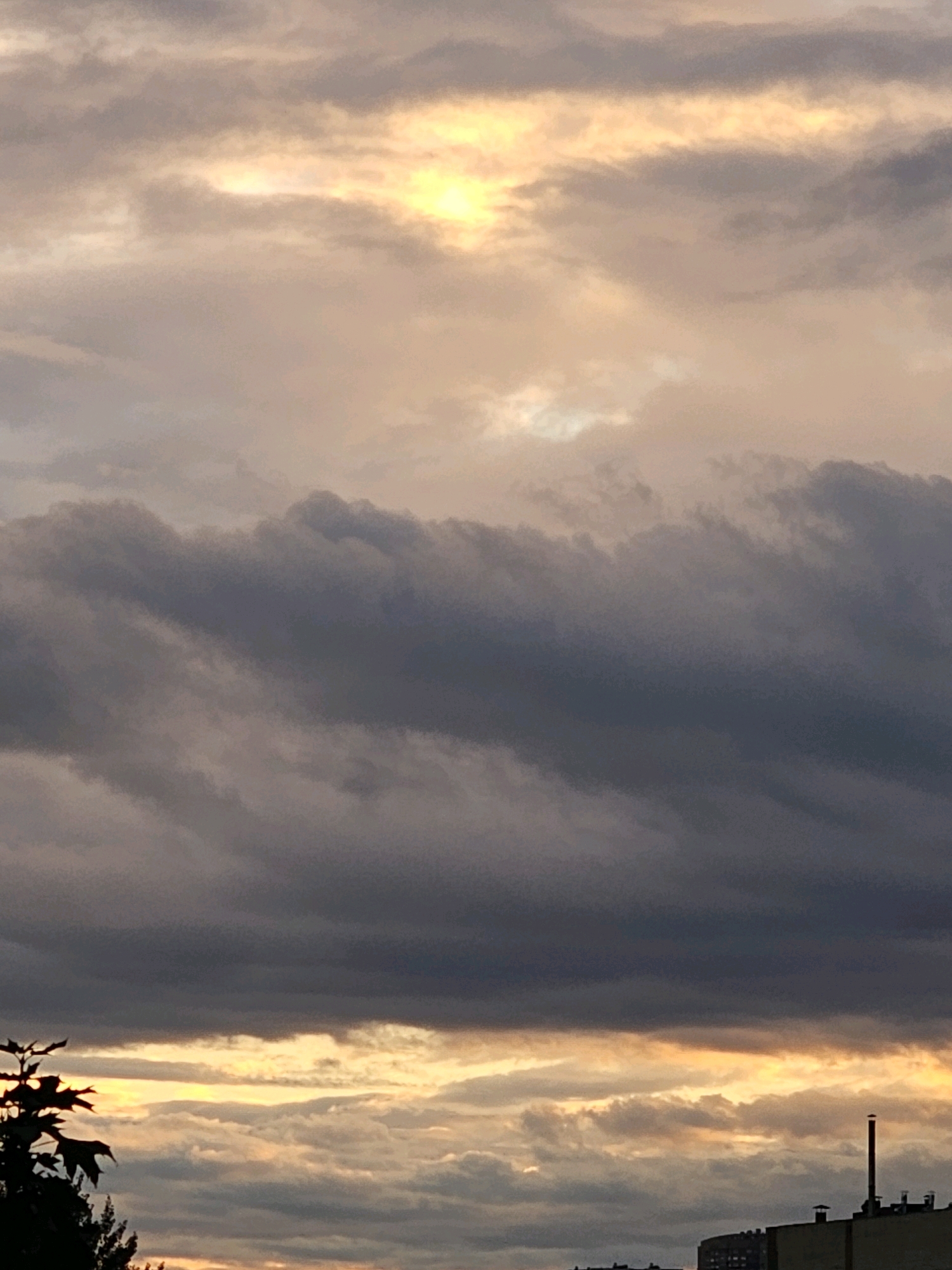 The image depicts a cloudy sky at dusk over the city of Khimki, Russia. In the foreground, silhouetted against the colorful sky, are the dark shapes of trees on the left and buildings and a smokestack on the right. The clouds are varied in color and texture, with areas of dark gray and lighter shades of orange and yellow where the sun is breaking through. The overall impression is that of an approaching evening or a storm, with dramatic lighting.