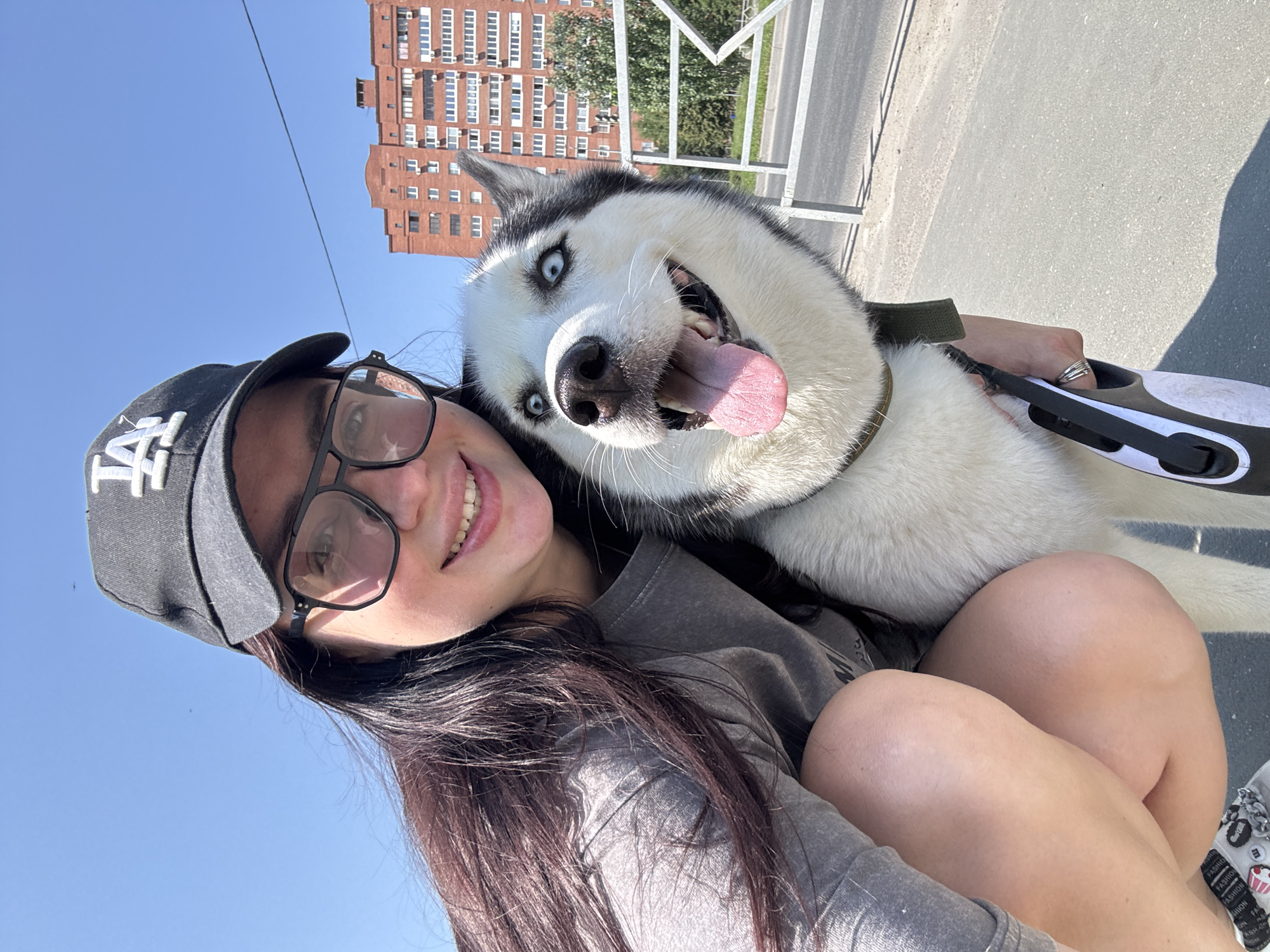 A selfie is captured outdoors on a clear day. A young woman, wearing glasses, a grey t-shirt, and a black baseball cap with a white logo, is crouching down. She has dark hair and is smiling. Next to her is a husky dog, which is predominantly white with black markings and striking blue eyes. The dog is looking directly at the camera with its tongue out. In the background, a tall, brick apartment building with many windows is visible, along with some trees and a white metal fence. The foreground shows a paved surface, likely a sidewalk or road. The location appears to be a residential area in the city of Tolyatti, Russia.