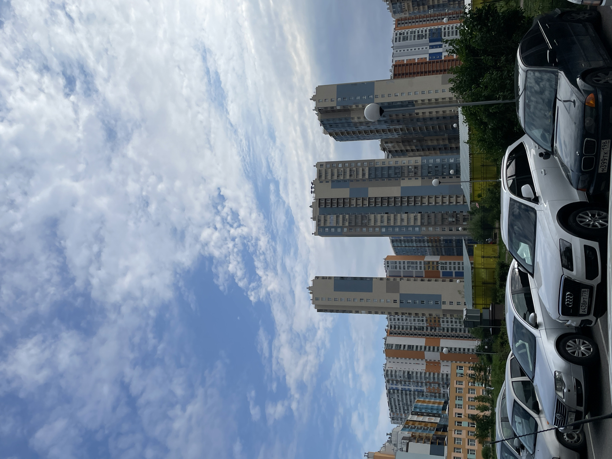 The image shows a city view of residential buildings and parked cars. The buildings are modern high-rises with colorful facades, characteristic of new urban developments. The sky is partly cloudy, with scattered white clouds against a blue background. In the foreground, several cars are parked in a lot, including a dark BMW, a white Audi SUV, and two silver sedans. The environment appears to be a residential district, possibly in the process of development or expansion, given the uniformity of the architecture. The overall impression is that of a typical urban landscape in a modern Russian city. The distinct architecture of the buildings strongly suggests the Kvartal u reki Orlovka residential complex in St. Petersburg, Russia.