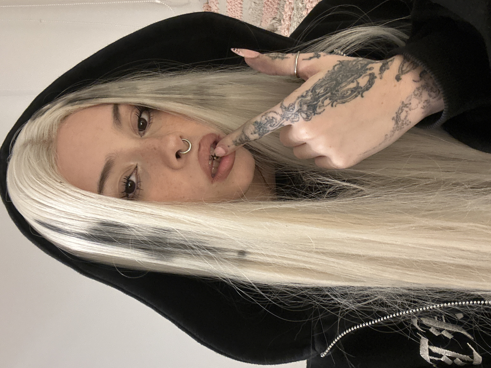 A portrait of a person with fair skin, light brown eyes, and bleached blonde hair wearing a black hooded sweatshirt. The person has a septum piercing and freckles are visible on their face. The person is holding their finger to their lower lip. The hand has several tattoos. The person appears to be inside. The image does not provide enough information to determine a specific location in Buenos Aires or offer forensic details.