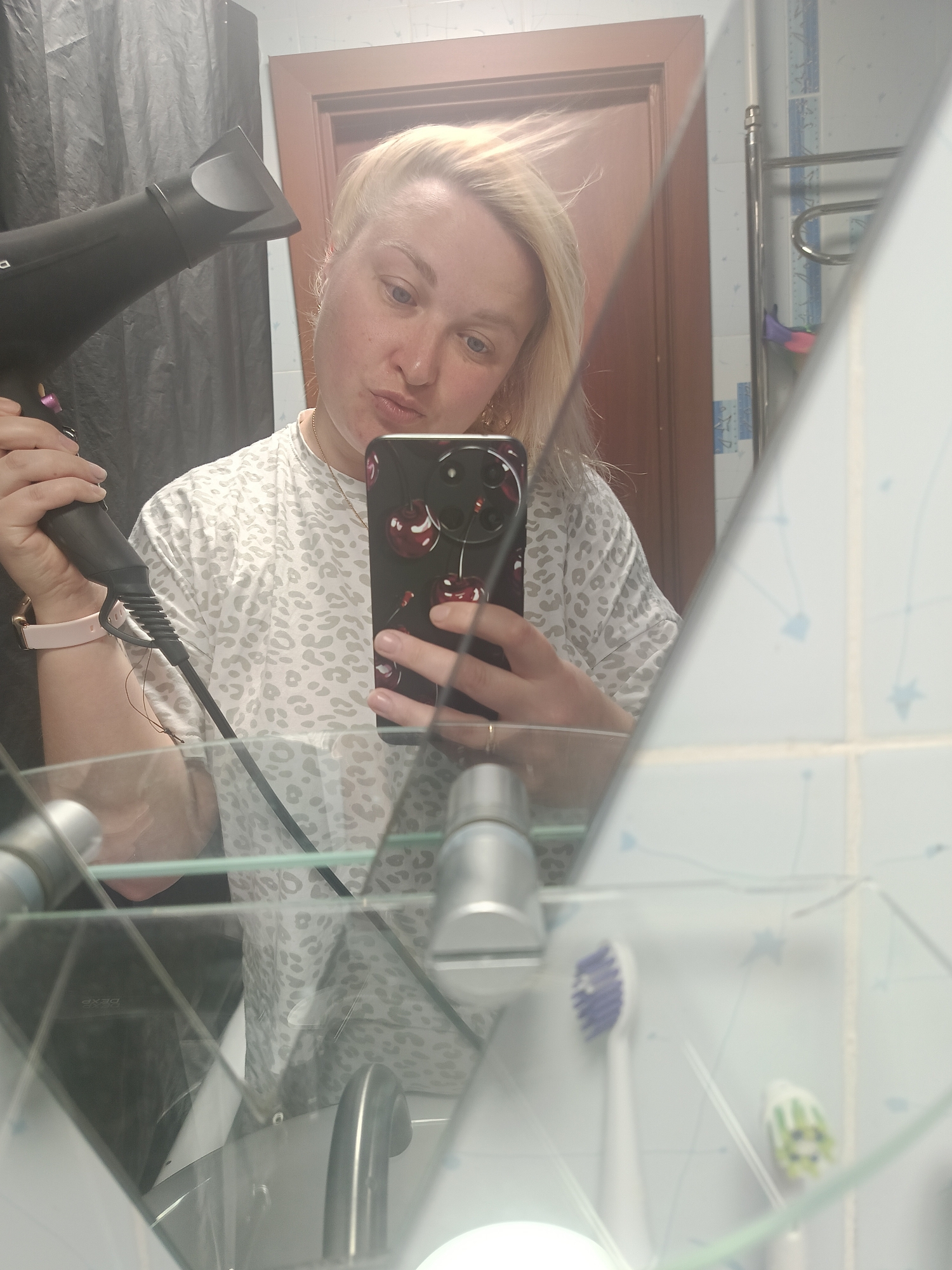 A woman is taking a selfie in a bathroom mirror. She is holding a hair dryer in her left hand and a phone with a cherry-themed case in her right hand. She has blonde hair and is wearing a white t-shirt with a leopard print pattern. The bathroom has light blue tiled walls. A door is visible in the background. Toothbrushes are visible in the reflection of the mirror, and a black curtain hangs on the left side of the scene.
The city is Chaikovsky, Russia.
