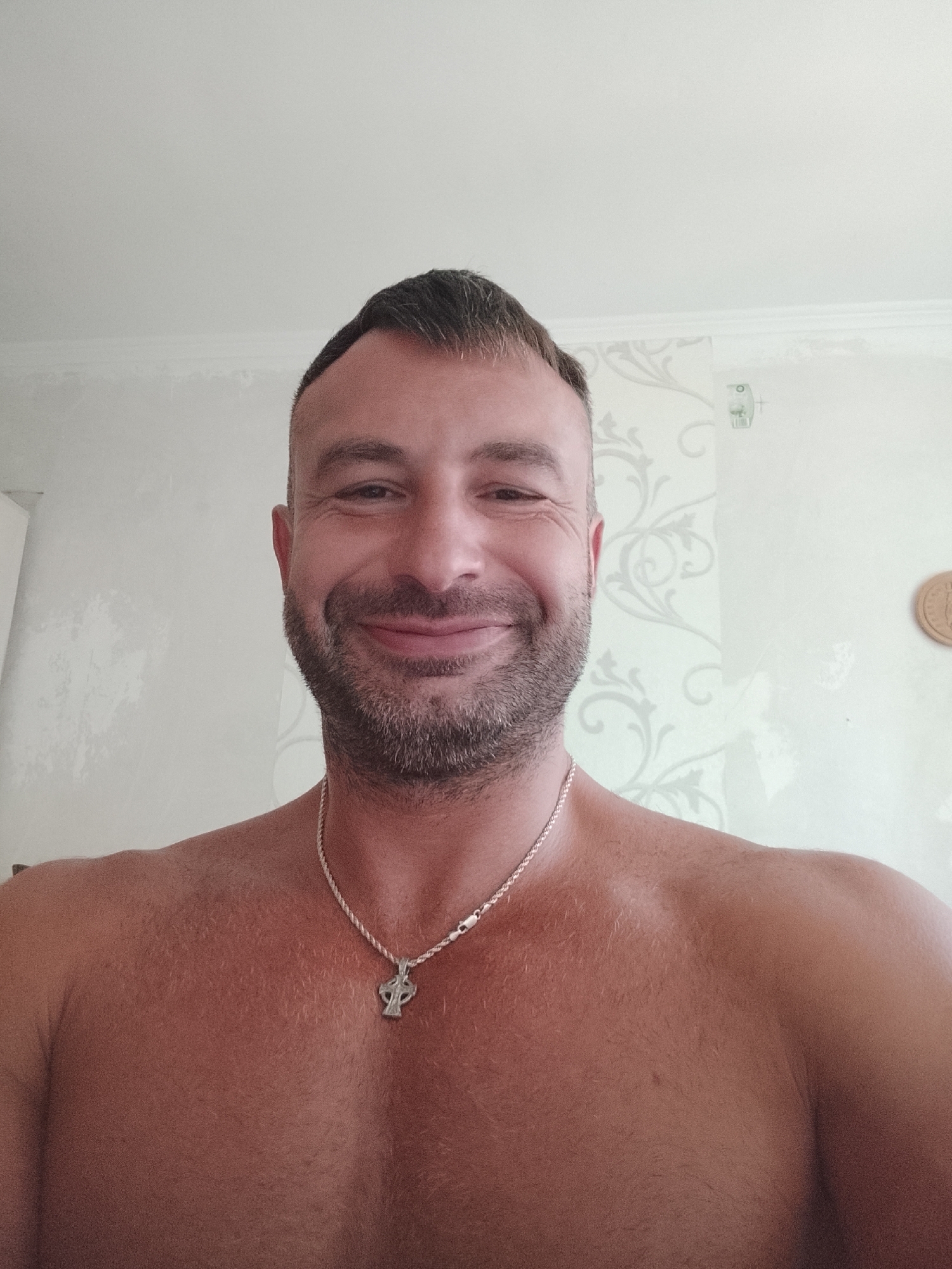 A shirtless man with short dark hair and a beard smiles at the camera. He is wearing a silver chain with a cross pendant. The background appears to be a wall with light-colored wallpaper featuring a floral pattern. The lighting is bright, suggesting it is daytime. There is no indication of the specific location within Chișinău, Moldova.
