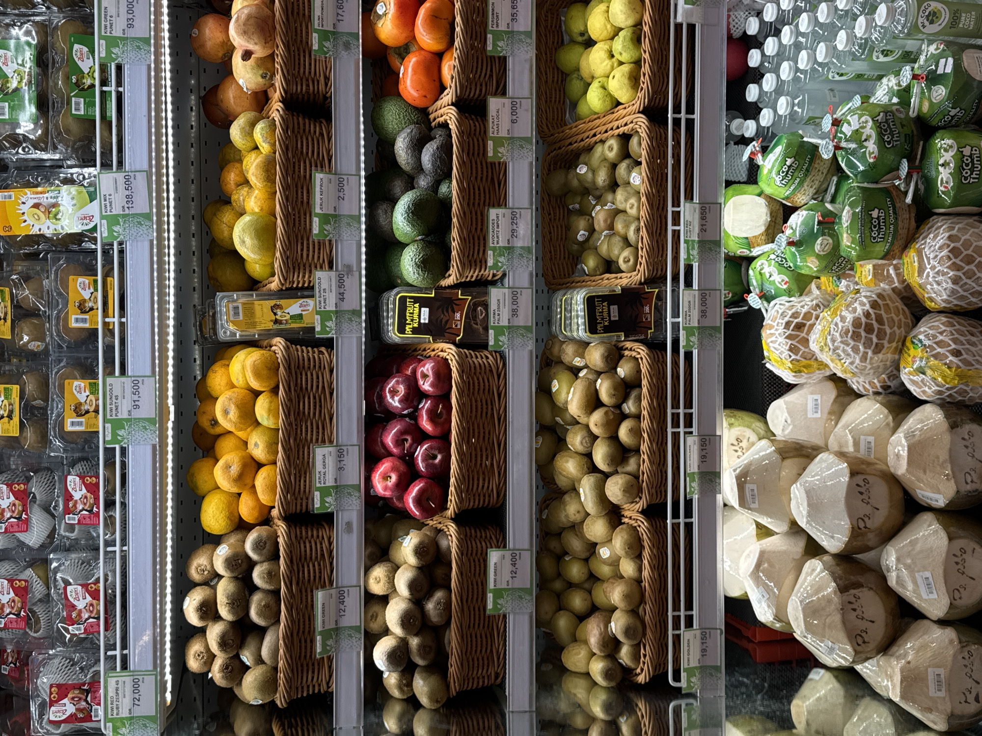 The image shows a refrigerated display shelf in a supermarket in North Kuta, Indonesia, filled with various fruits. On the top shelf, packaged kiwi fruits are visible. The middle shelves contain baskets of fruits, including kiwi, oranges, apples, avocados, persimmons, and what appear to be pomegranates or similar round red fruits. Some baskets are labeled with product names and prices. The bottom shelves display more fruits in baskets, including kiwi and what seem to be guavas or pears. In the foreground, there are numerous coconuts, some wrapped in netting and others with their tops covered, and several bottles of coconut water. The overall environment appears to be a clean and well-stocked produce section of a retail store. There are no people visible in the image.