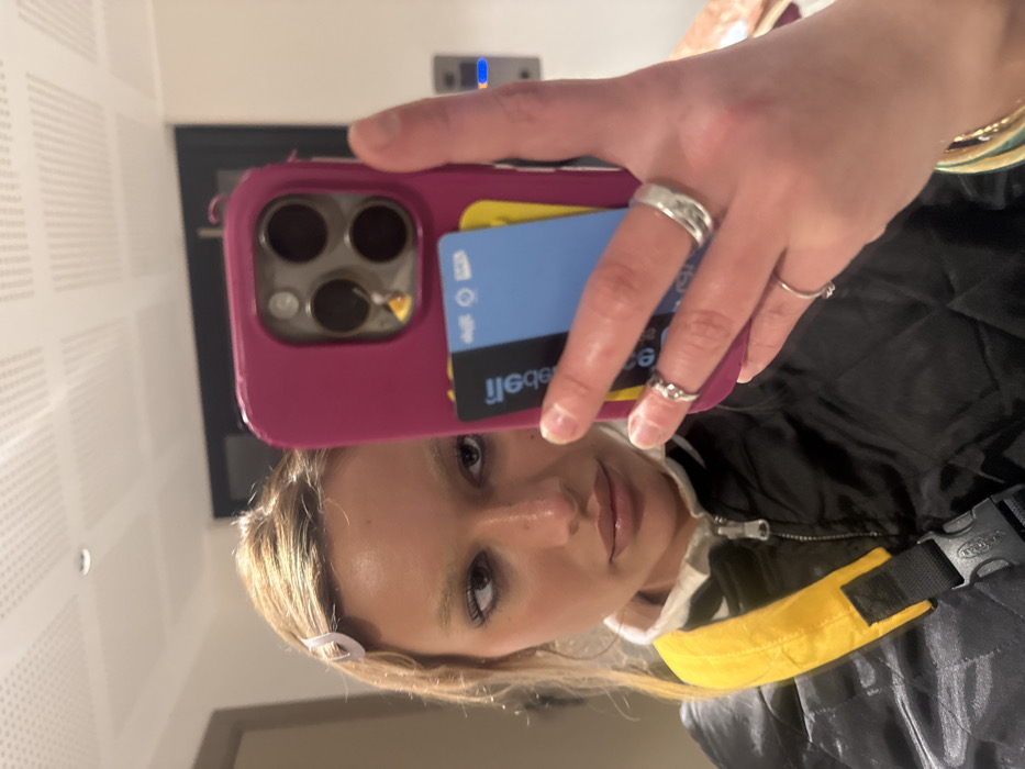 A person takes a selfie in front of a wall. They are holding a pink phone case with a blue credit card and a black transit card tucked into the back. The person has several rings on their fingers. They are wearing a black jacket with a yellow strap. The image is taken at an angle, and the person's face is tilted.