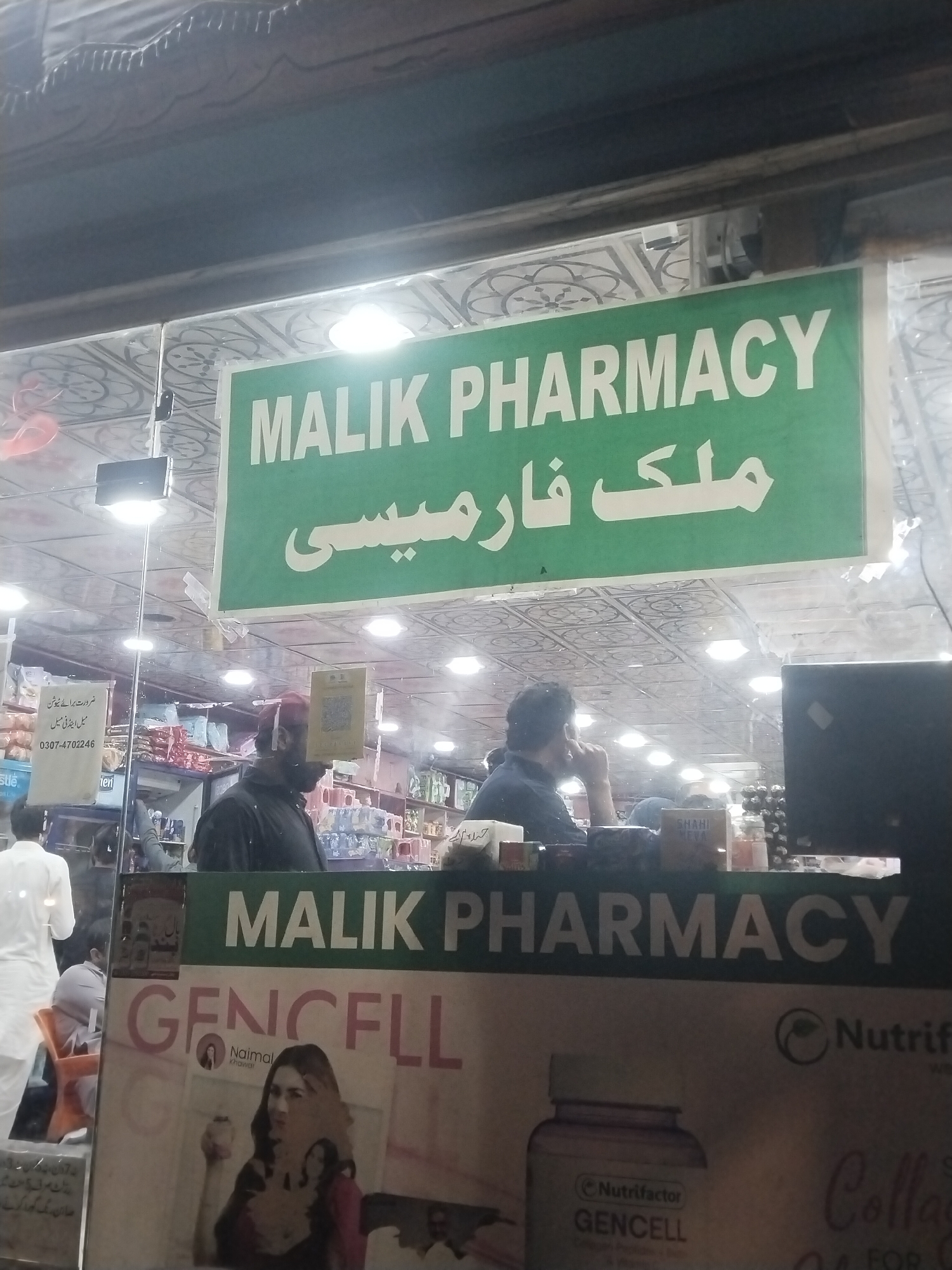 The image depicts the front of Malik Pharmacy in what appears to be a city in Pakistan, possibly Heir. The pharmacy's name is displayed in both English and Urdu on a green sign. The interior is visible through the storefront window, with people present, including a man with a red cap. There is a poster advertising "Gencell" with a woman on it. Also visible are advertisements for tutoring services, featuring the phone number 0307-4702246. A variety of products can be seen on the shelves inside the pharmacy. The lighting suggests it is evening or night.