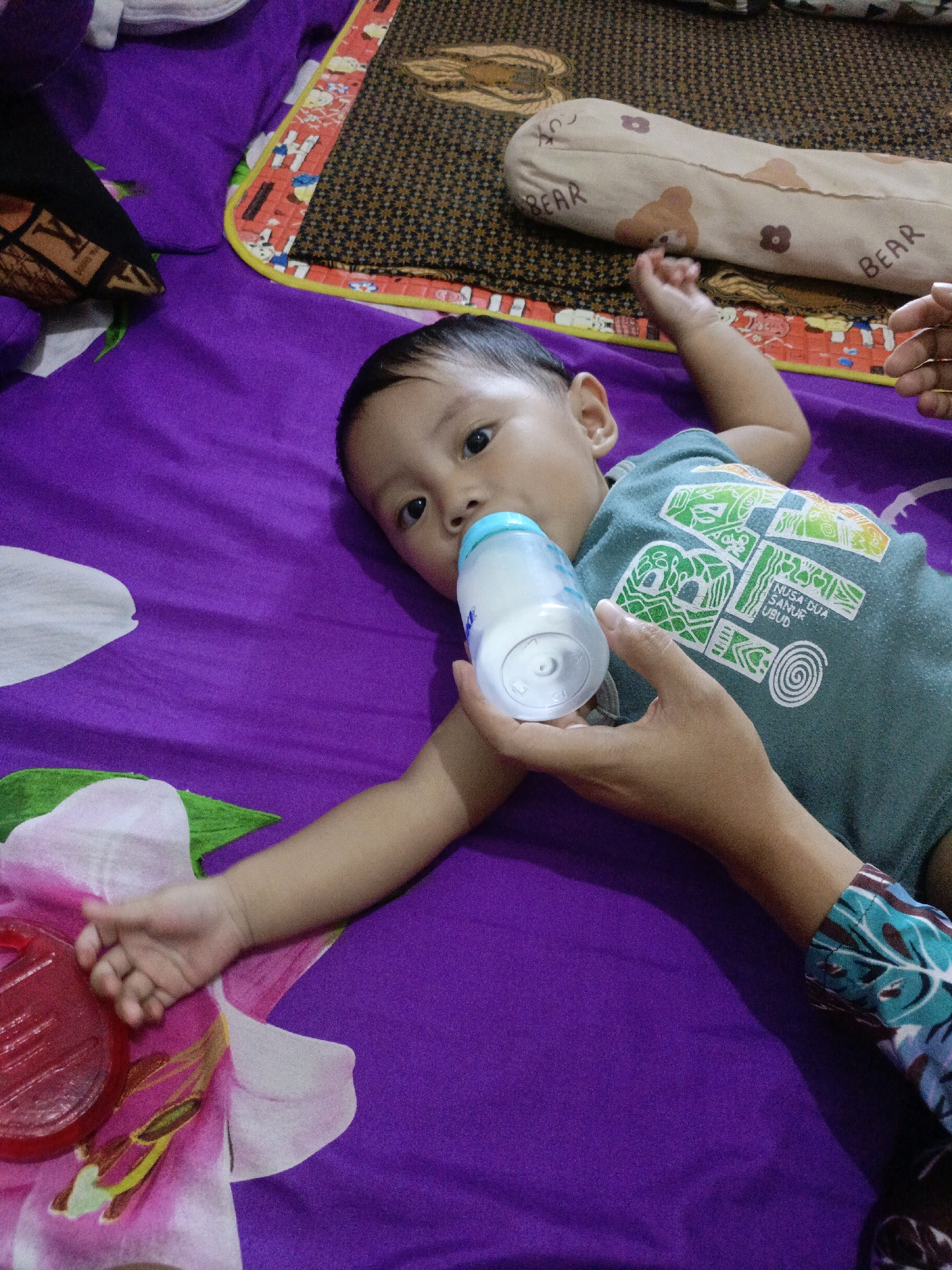 A baby is lying on a purple bedsheet and drinking from a milk bottle held by a person's hand. The baby is wearing a green t-shirt with a graphic design. A patterned mat with a bear-shaped pillow is visible behind the baby. The scene is likely indoors, possibly in a bedroom in Pager, Indonesia.